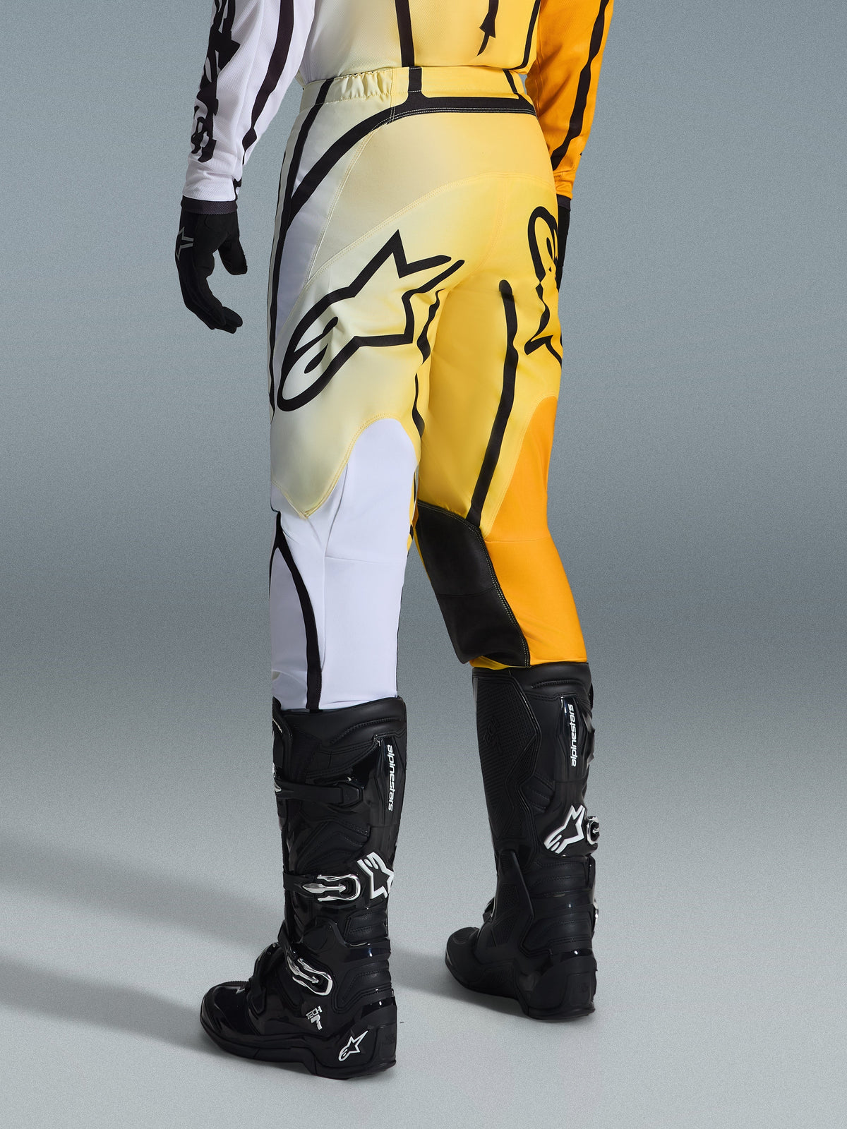 Alpinestars 2026 Fluid Apex Pants, calças de motocross, degradê amarelo e preto com detalhes brancos e cor de laranja, vista traseira, usadas com camisola a condizer e botas Tech 7 pretas, com um logótipo Alpinestars preto de destaque no assento, equipamento de performance para condução fora de estrada