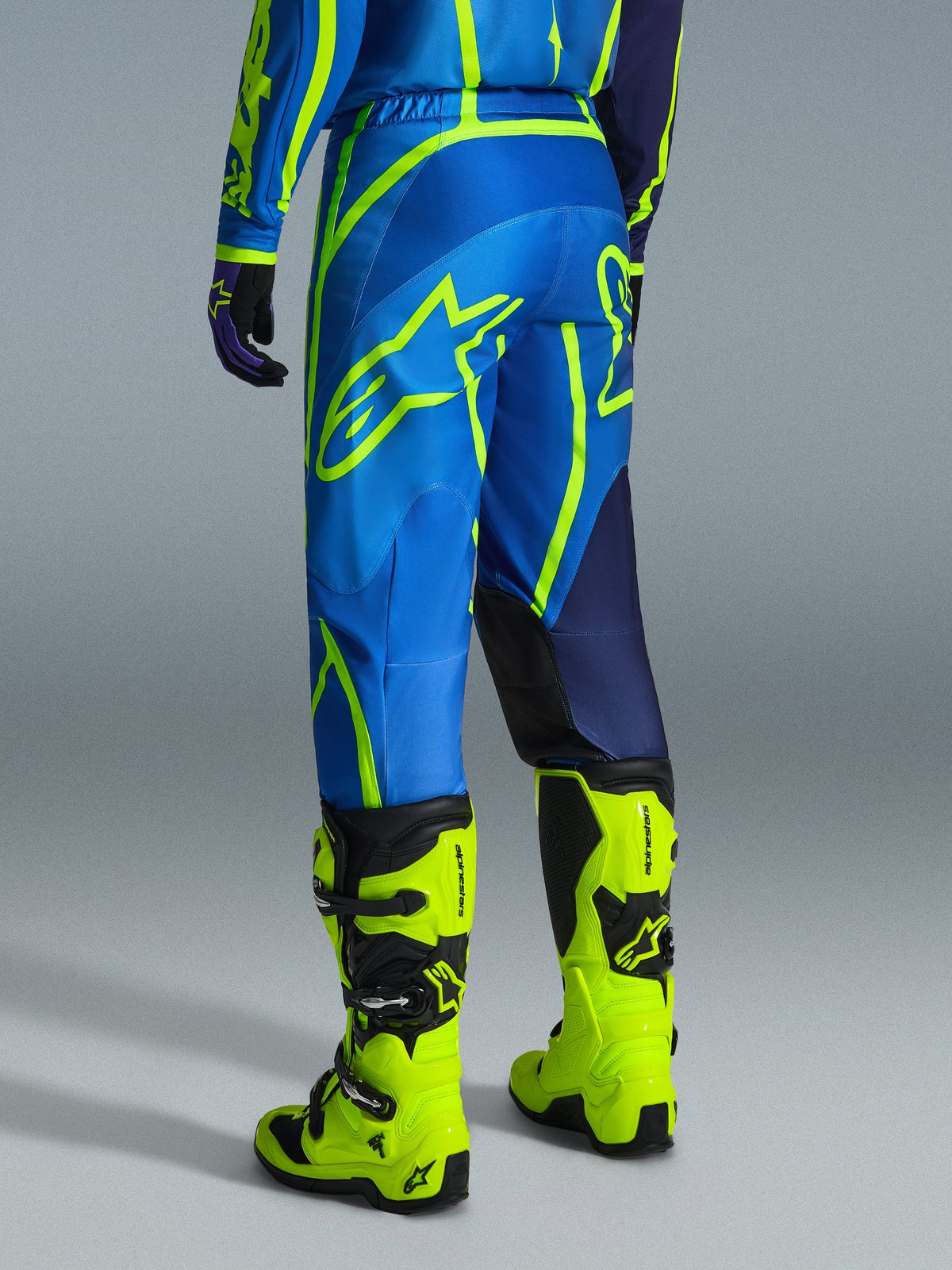 Alpinestars 2026 Fluid Apex Pants, calças de motocross, azul e amarelo néon, vista traseira do piloto a usar equipamento a condizer e botas amarelo fluorescente, com logótipos Alpinestars marcantes e design ergonómico