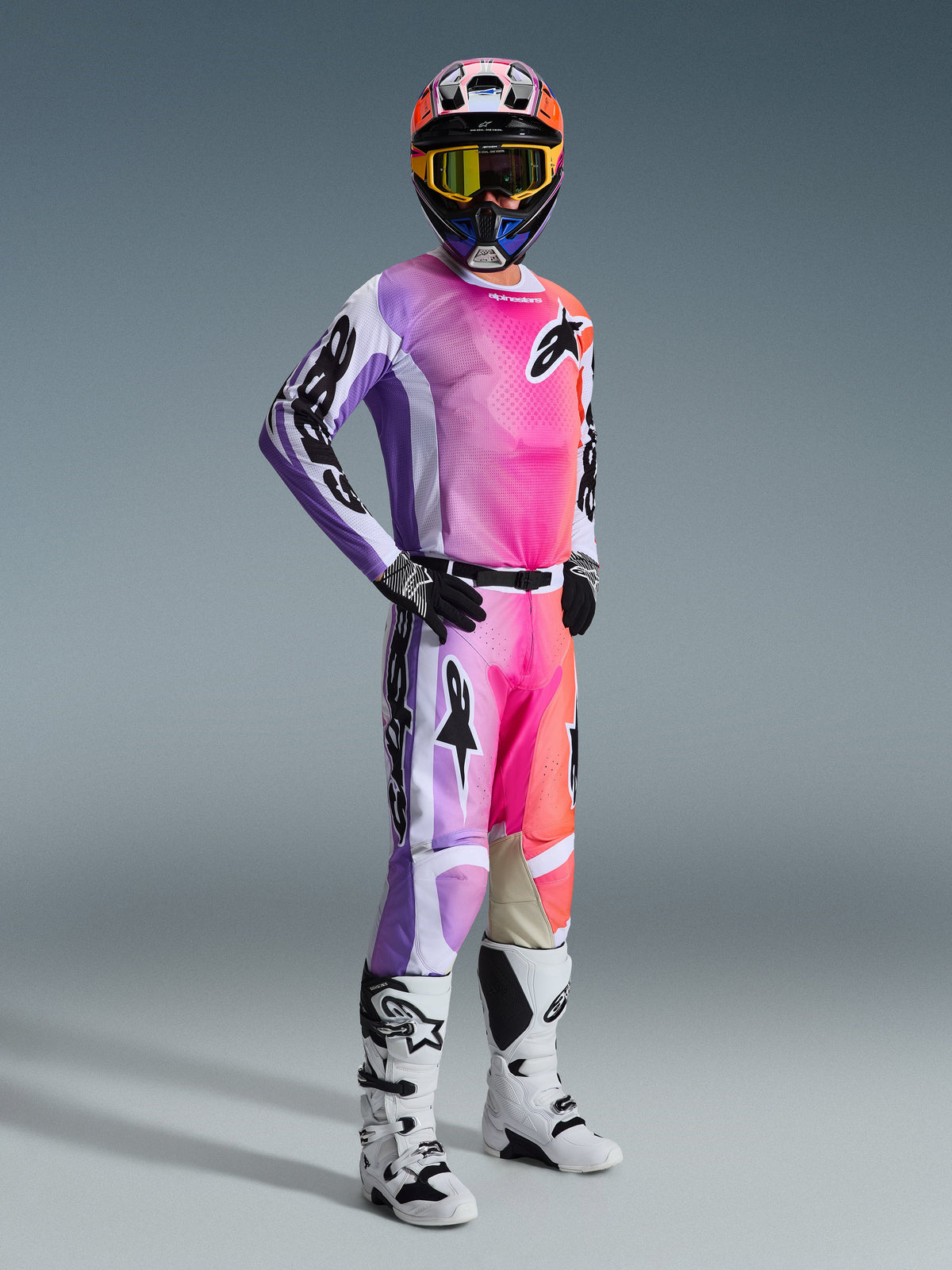 Motocross rider wearing the Alpinestars 2026 Racer Air Portl Jersey and matching pants, vibrant pink, purple, and orange multicolor gradient with white accents, coordinated colorful helmet and yellow goggles, white and black boots, complete racing outfit for off-road performance