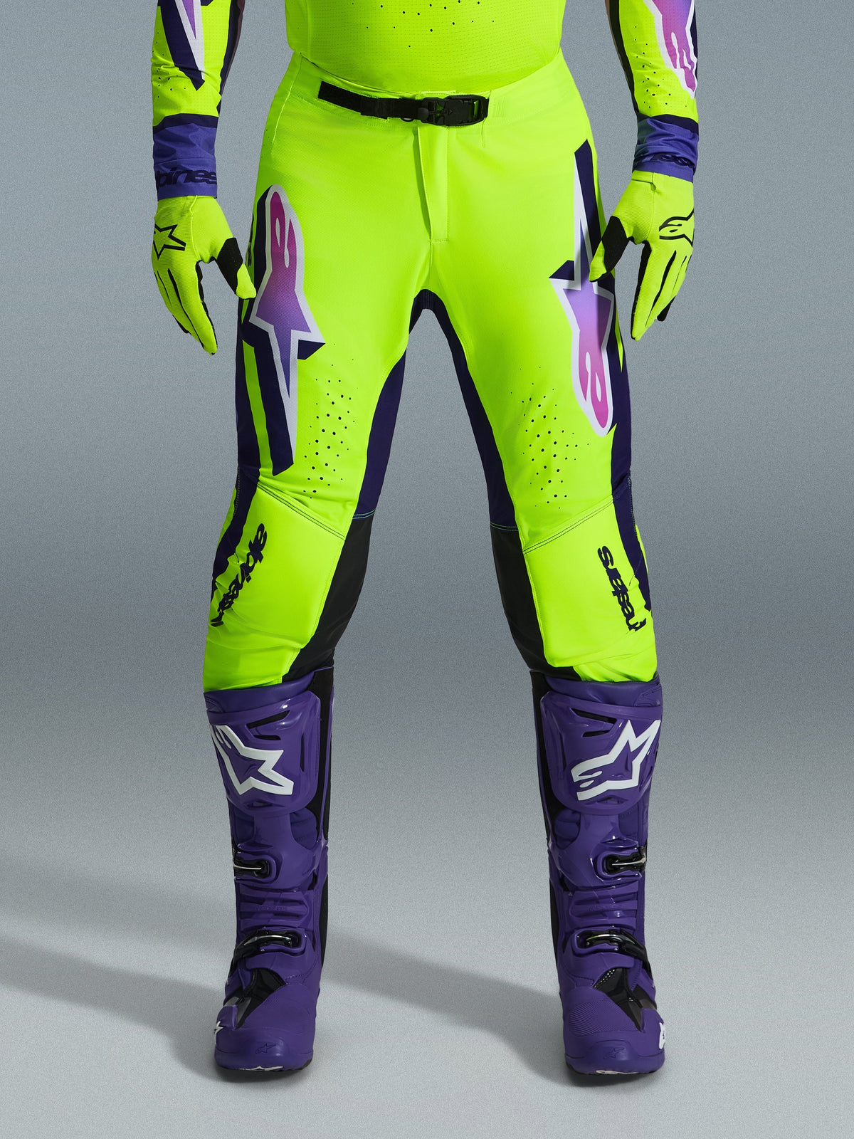 Alpinestars 2026 Supertech Vista Pants, calças de motocross, amarelo fluorescente, roxo e preto, combinadas com luvas a condizer e botas roxas, com grandes logótipos em degradê nas coxas, zonas perfuradas para ventilação e design atlético profissional para corridas todo-o-terreno