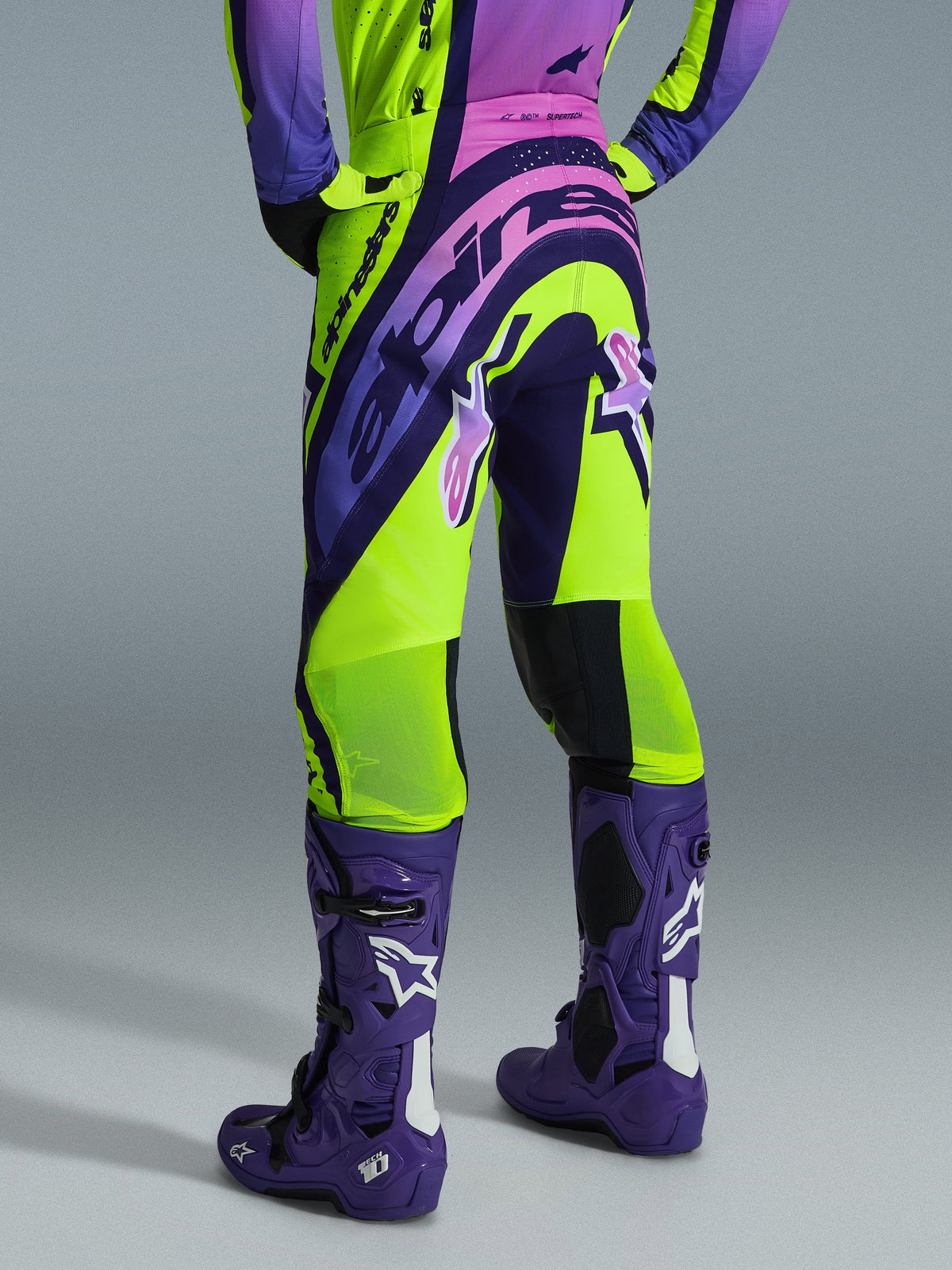Alpinestars 2026 Supertech Vista Pants, calças de motocross, amarelas, roxas e pretas, vista traseira com camisola a condizer e botas de motocross roxas, com logótipos em destaque e zonas de malha respirável nas panturrilhas