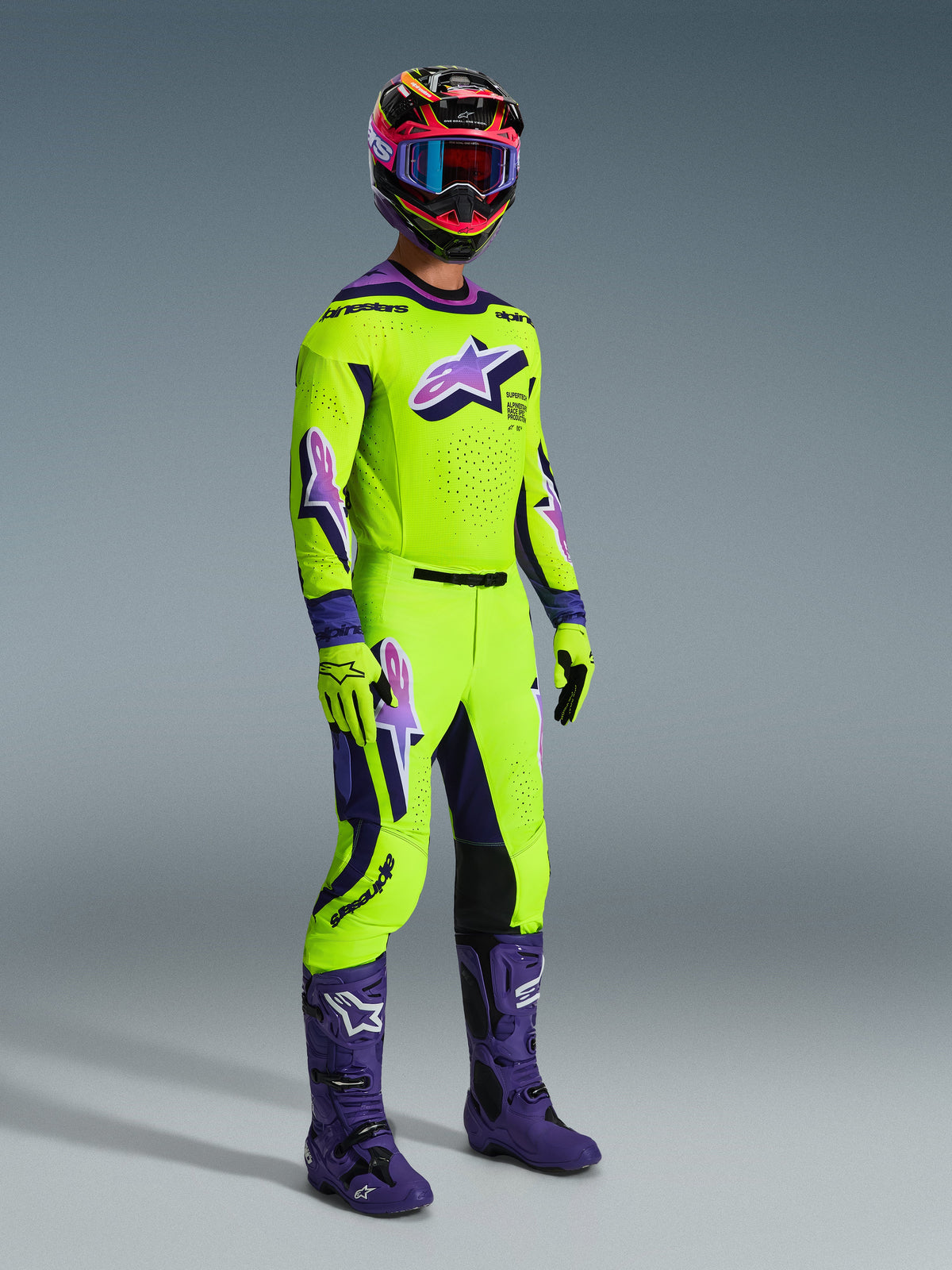 Alpinestars 2026 Supertech Vista Gear Set, professional motocross rider wearing a vibrant lime green jersey and pants with purple accents and logos, matching lime green gloves, purple boots, and a multi-colored helmet with blue goggles, coordinated athletic-fit gear for off-road racing