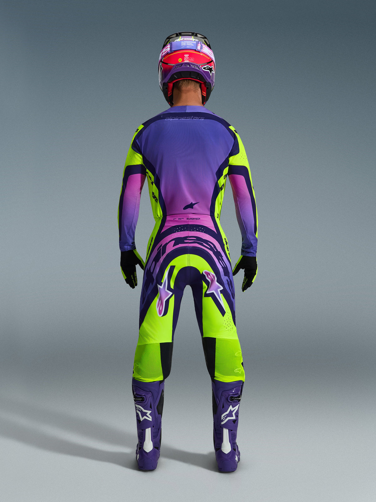 Alpinestars 2026 Supertech Vista Gear Set, back view of a motocross rider wearing a vibrant purple, pink, and lime green gradient racing outfit, featuring matching jersey and pants with bold branding, coordinated purple and pink helmet, purple boots, and black gloves.
