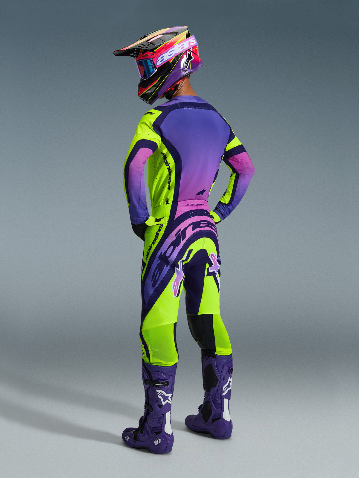 Motocross rider from the back wearing Alpinestars 2026 Supertech Vista Gear Set, purple jersey with pink gradient and neon yellow accents, matching neon yellow and purple pants, deep purple boots with white logos, coordinated helmet and goggles, pro-level motocross racing apparel