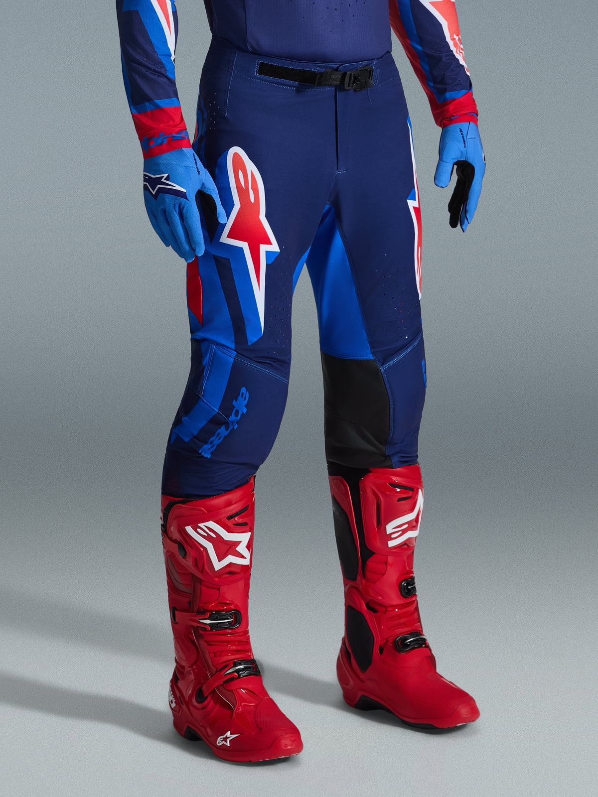 Alpinestars 2026 Supertech Vista Pants, calças de motocross, Dark Navy Hot Coral Cobalt, usadas com botas de corrida em vermelho vivo e luvas azuis, com logótipos gráficos grandes nas coxas e painéis internos reforçados pretos nos joelhos para performance todo-o-terreno.