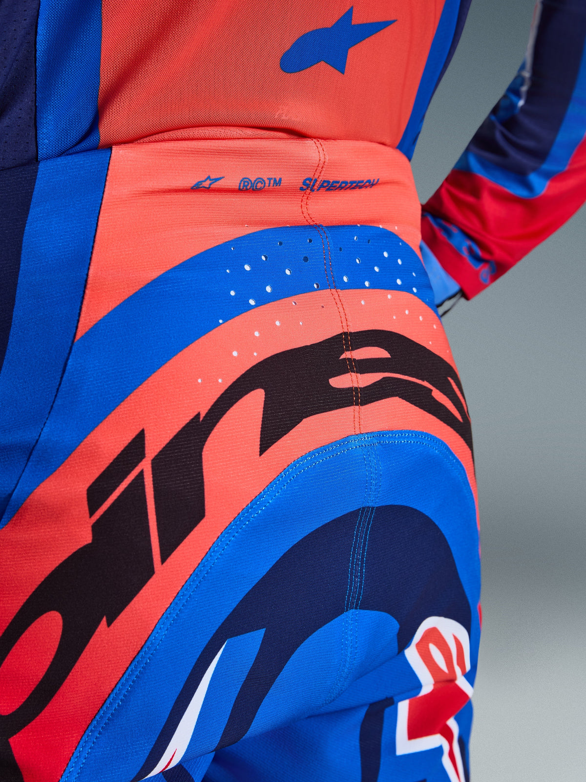 Pormenor das Calças Alpinestars 2026 Supertech Vista, calças de motocross, em Hot Coral e Cobalt Blue, com um grande grafismo Alpinestars a preto no assento, furos de ventilação cortados a laser e marca 'SUPERTECH' na parte de trás da cintura, vestuário de competição de nível profissional