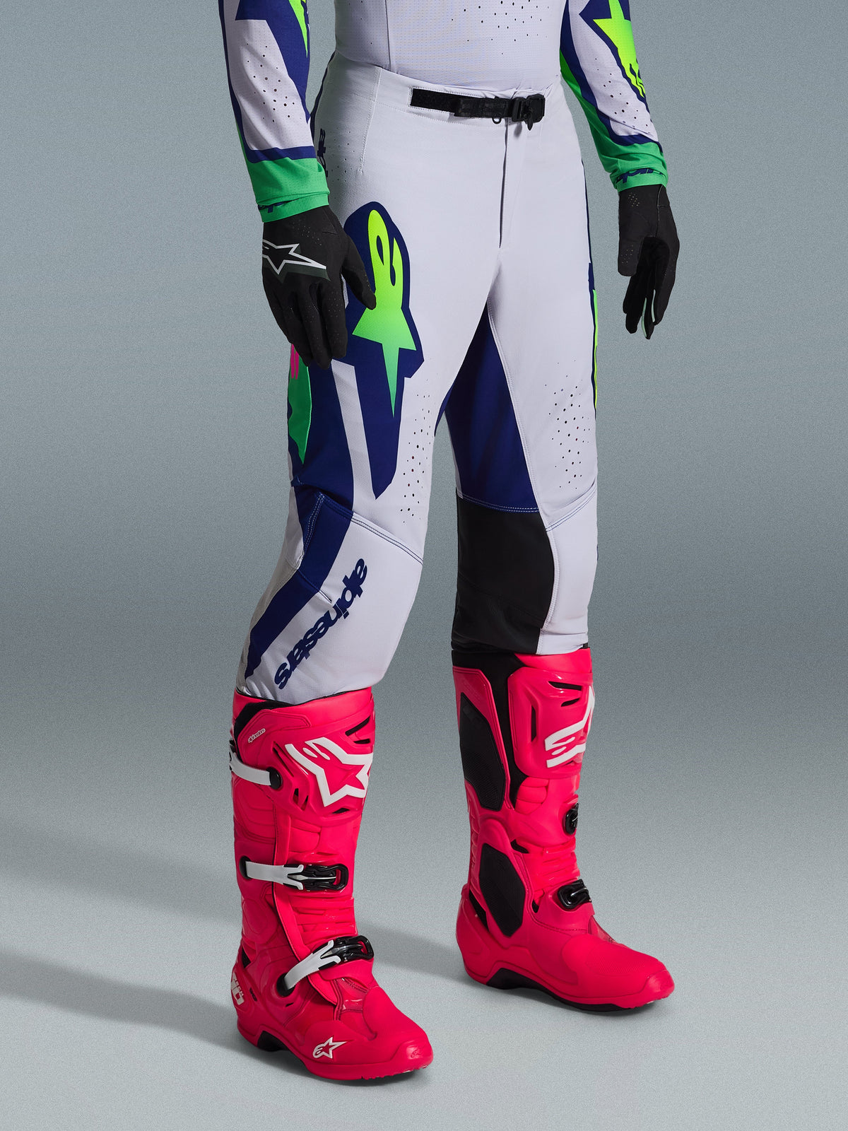Alpinestars 2026 Supertech Vista Gear Set, calças de motocross, cinzento claro com detalhes em azul-marinho e verde-lima, usadas com botas rosa vibrante e luvas pretas, apresentando um corte atlético profissional e grafismos arrojados