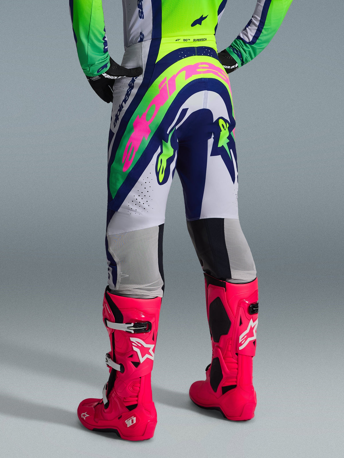 Calças Alpinestars 2026 Supertech Vista, calças de motocross, cinzento claro, amarelo fluo e azul, vista traseira com um grande logótipo Alpinestars cor-de-rosa, usadas com camisola de corrida a condizer e botas Tech 10 cor-de-rosa vibrante, equipamento profissional de todo-o-terreno