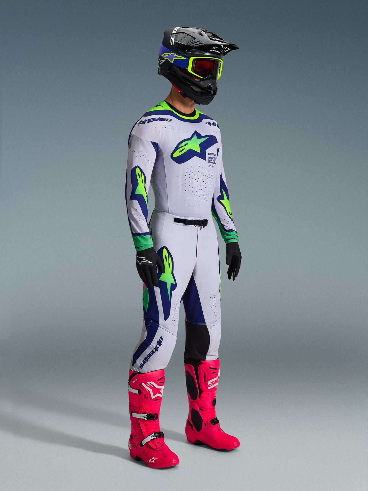 Alpinestars 2026 Supertech Vista Gear Set, piloto de motocross com equipamento de corrida completo, camisola e calças brancas com detalhes em verde lima e azul marinho, botas cor-de-rosa vibrante, capacete preto com detalhes em azul e lima, vestuário de todo-o-terreno coordenado de nível profissional