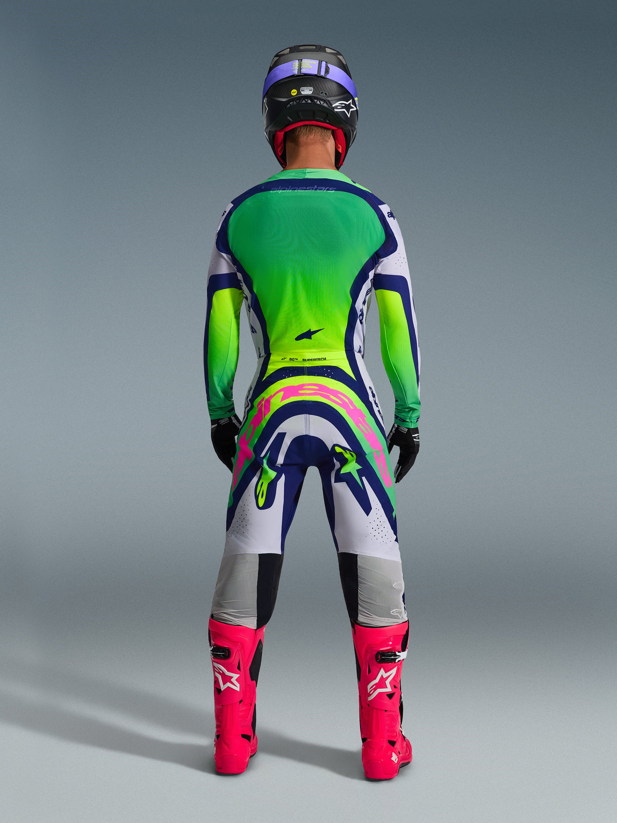 Conjunto de Equipamento Alpinestars Supertech Vista 2026, vista traseira de conjunto de motocross, camisola com gradiente em verde e amarelo, calças brancas e azuis com um grande logótipo cor-de-rosa na parte traseira, botas cor-de-rosa vibrante e capacete roxo, vestuário profissional para todo-o-terreno