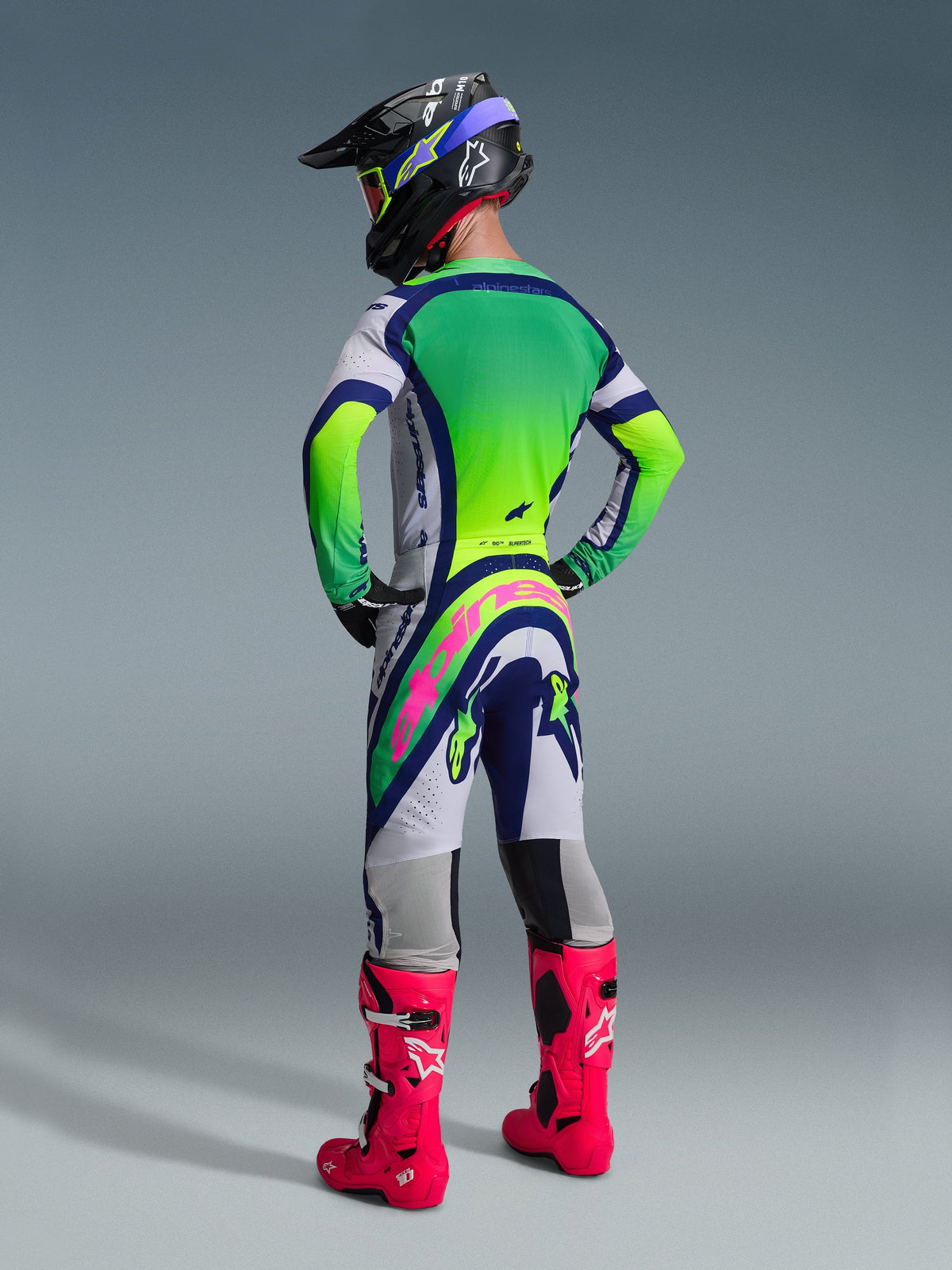 Piloto de motocross visto de trás a usar as calças Alpinestars 2026 Supertech Vista, com design em cinzento claro, azul e amarelo néon e um logótipo cor-de-rosa vibrante no assento, combinadas com uma camisola a condizer, capacete preto e botas cor-de-rosa vibrante para um kit completo de corrida todo-o-terreno.