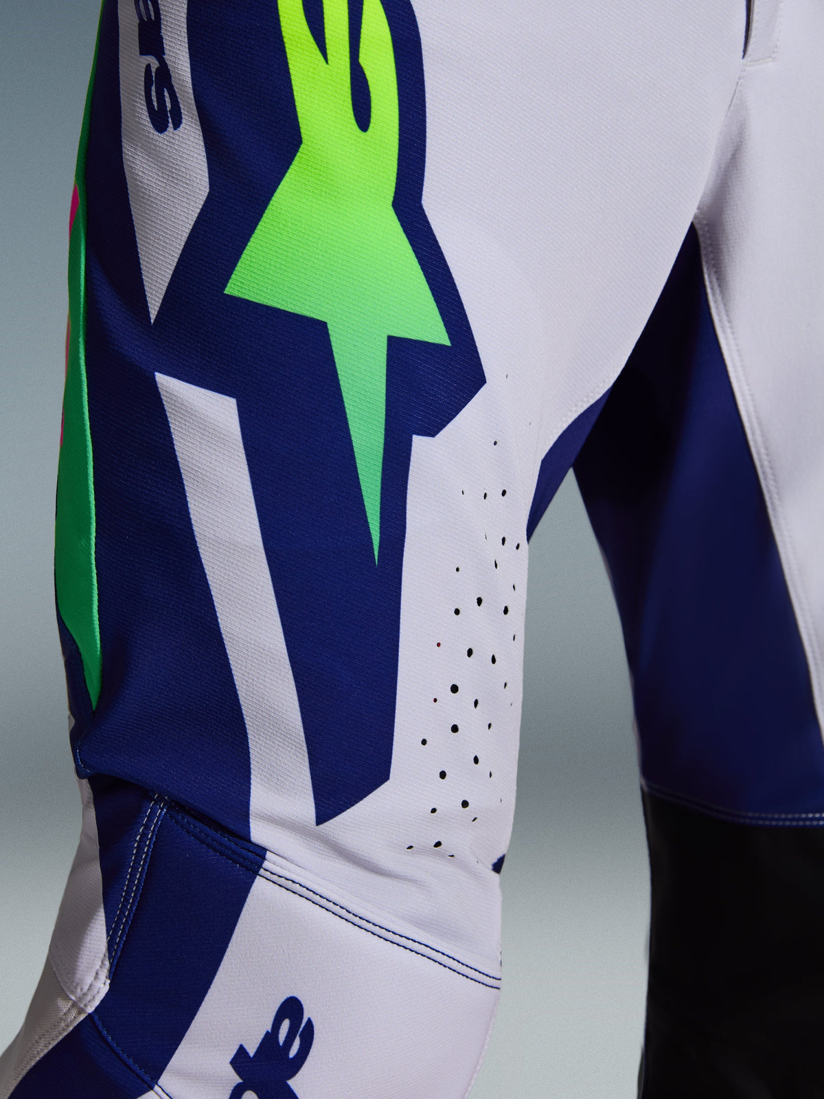 Calças Alpinestars Supertech Vista 2026, calças de motocross, cinzento claro com detalhes em amarelo fluo e azul, vista aproximada da coxa mostrando o logótipo A-star de grandes dimensões, orifícios de ventilação cortados a laser e tecido rip-stop elástico avançado