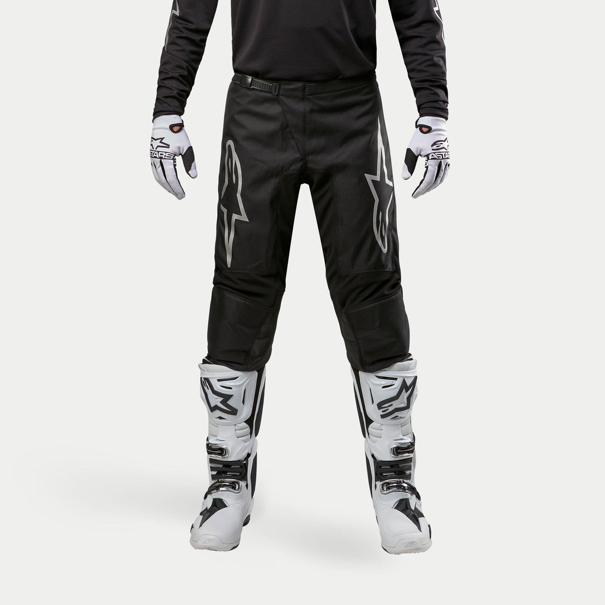 Alpinestars 2024 Fluid Graphite Pants, calças de motocross, pretas e prateadas, com logótipos Alpinestars prateados nas coxas, usadas com botas e luvas de motocross brancas e pretas, design desportivo para corridas todo-o-terreno