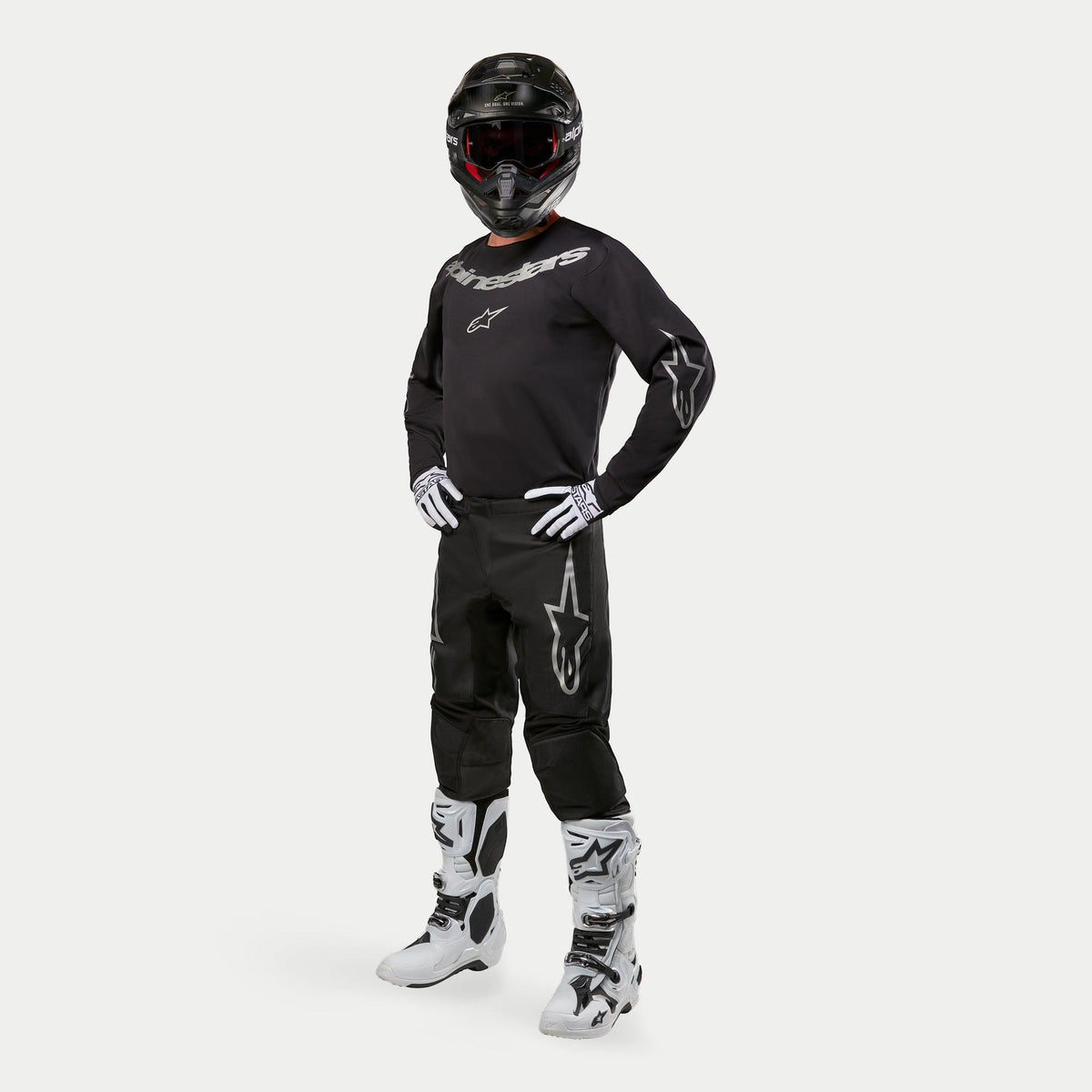 Calças Alpinestars 2024 Fluid Graphite, calças de motocross, pretas e prateadas, usadas com camisola preta a condizer, capacete preto, luvas brancas e botas brancas, com logótipos Alpinestars prateados nas coxas, ajuste atlético ergonómico para corridas todo-o-terreno