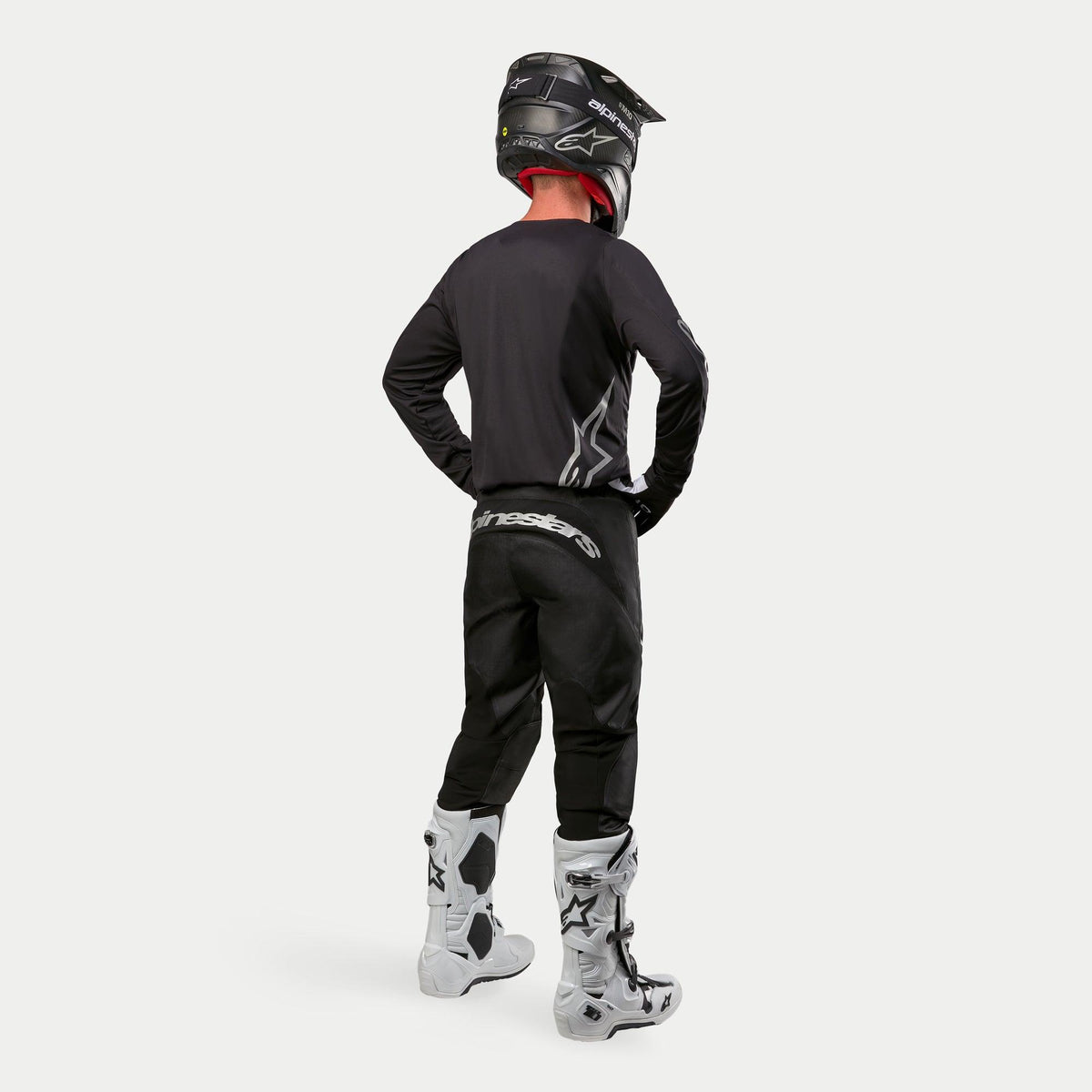 Alpinestars 2024 Fluid Graphite Pants, calças de motocross, preto e prateado, vista traseira de um piloto a usar camisola preta, capacete preto e botas brancas e pretas, com marca prateada na parte inferior das costas e construção durável em poli-tecido para corridas de todo-o-terreno