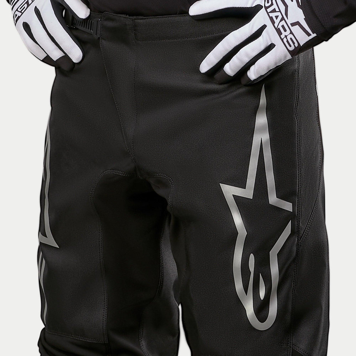 Alpinestars 2024 Fluid Graphite Pants, calças de motocross, pretas e prateadas, pormenor da zona superior das coxas e cintura, com um grande logótipo de estrela prateado na perna, usadas com luvas de corrida pretas e brancas