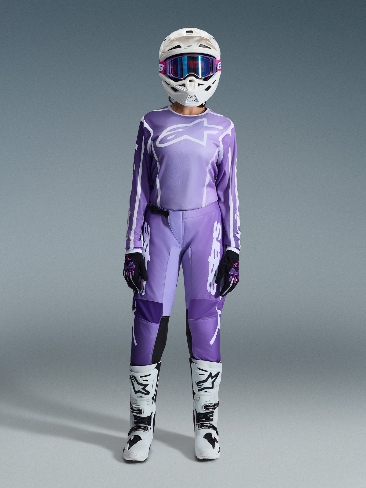 Motocross rider wearing the Alpinestars 2026 Woman Stella Fluid Apex Gear Set, violet and white jersey with matching pants, white helmet with pink goggles, and white and black off-road boots, complete racing kit for women's motocross.