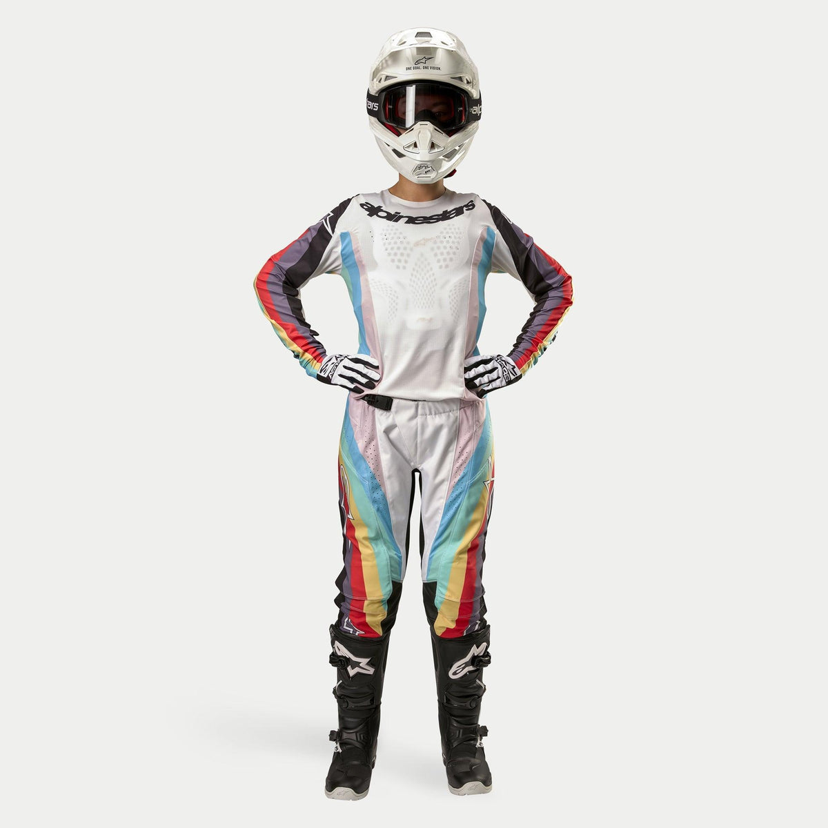 Female motocross rider wearing Alpinestars Women Stella 2024 Techstar Pants, Black Multicolor featuring white base with red, yellow, and blue stripes, matching long-sleeve jersey, white helmet, and black boots, full racing gear for off-road competition