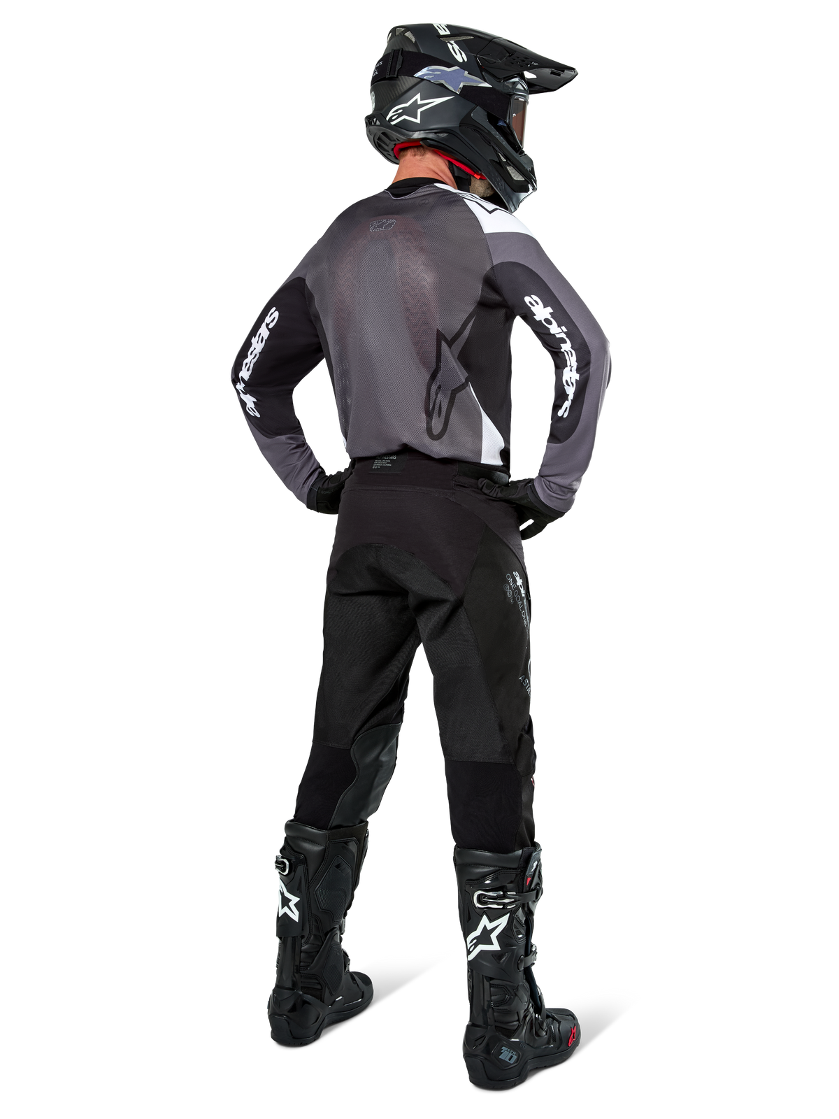 Piloto de motocross com camisola Alpinestars Pro-Dura em Black Magnet White, vista traseira mostrando painel traseiro em malha cinzenta e mangas pretas com marca Alpinestars branca, coordenada com calças pretas, capacete preto e botas pretas e brancas, equipamento profissional de enduro