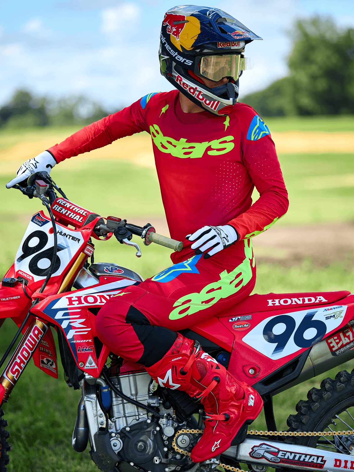 Piloto de motocross com a Camisola Alpinestars Supertech Maker 2025, em vermelho vivo com branding 'astars' em amarelo néon, combinada com calças e botas vermelhas a condizer, luvas brancas e pretas, sentado numa mota de corrida Honda vermelha num cenário ao ar livre