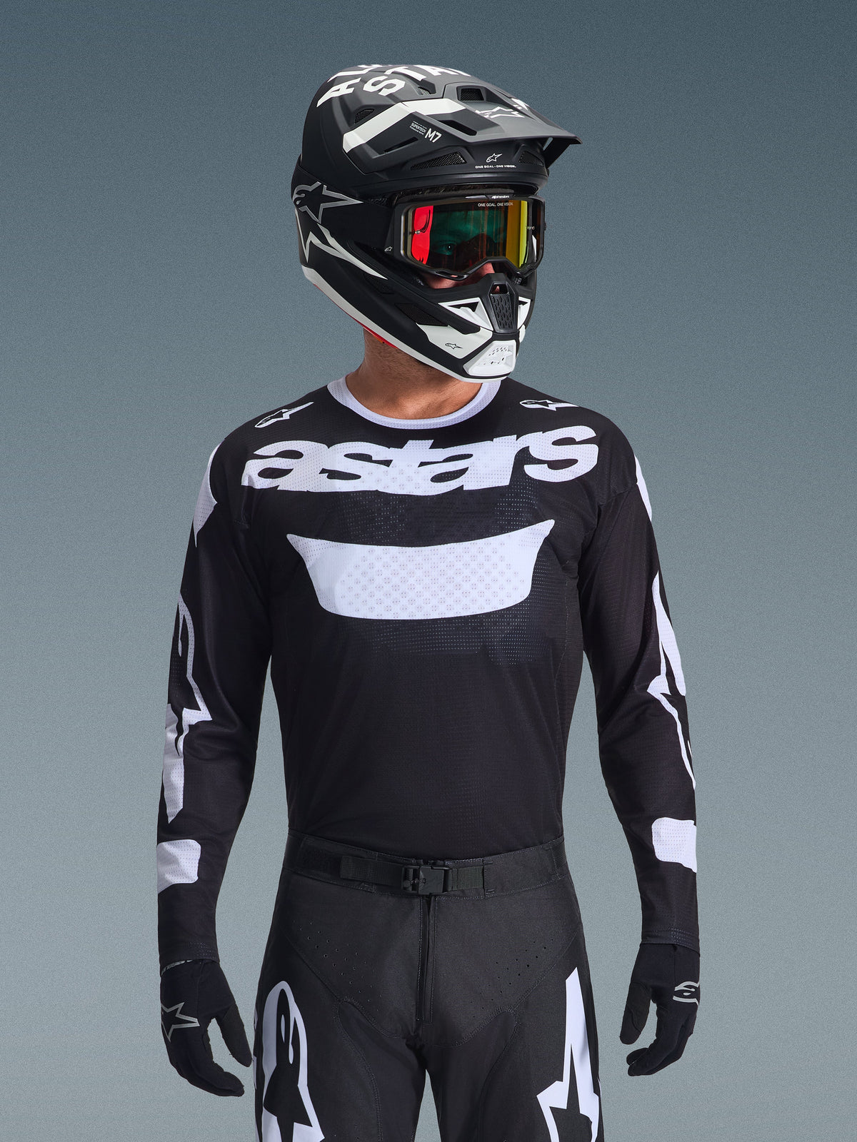 Piloto de motocross com o conjunto Alpinestars 2026 Racer Air Riway, camisola preta com logótipo 'astars' branco e painel ventilado no peito, calças pretas com detalhes brancos, capacete preto e branco a condizer com óculos iridescentes, luvas pretas, equipamento de motocross de alta performance em preto e branco.