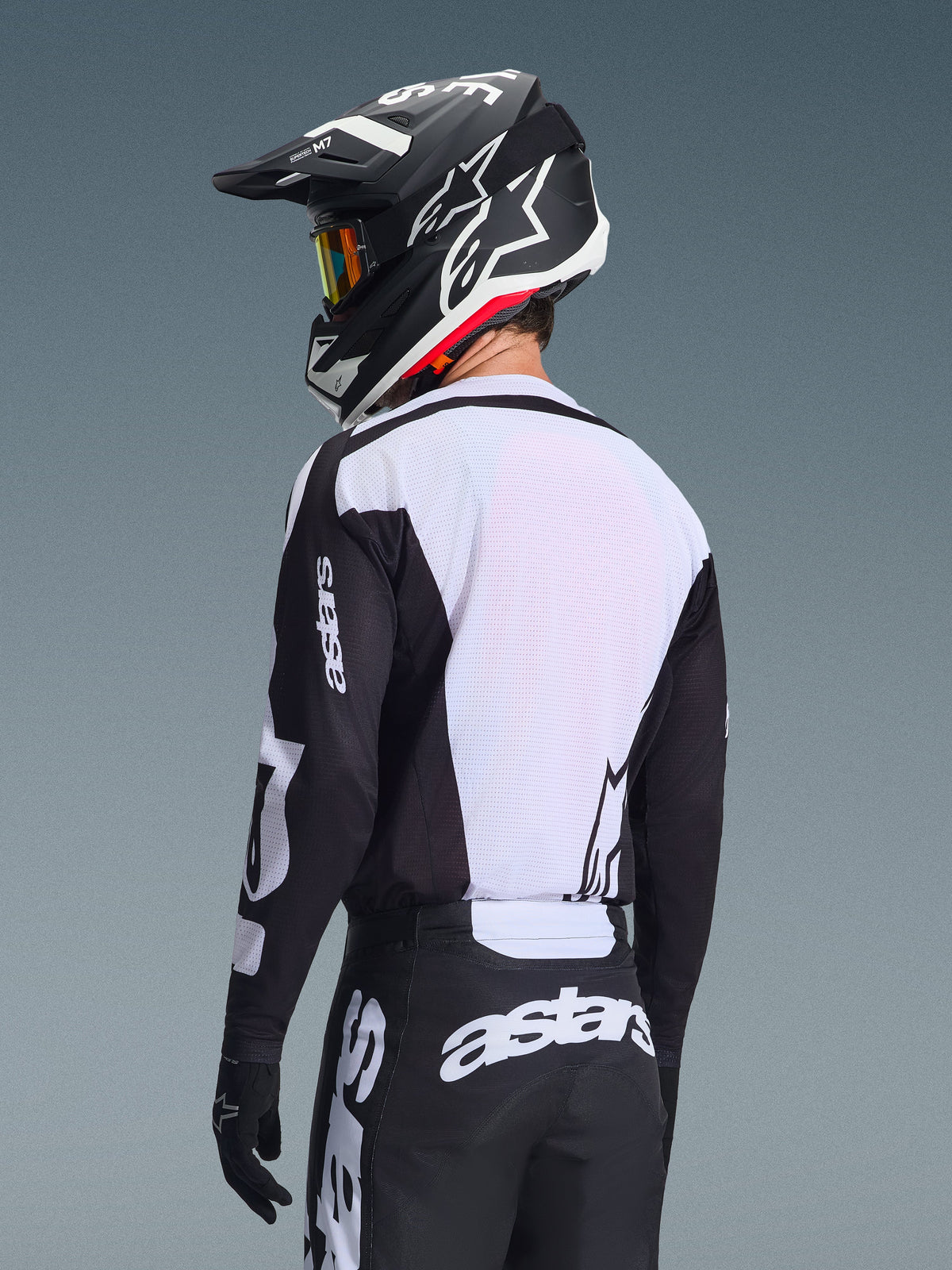 Piloto de motocross visto de costas a usar a camisola Alpinestars 2026 Racer Air Riway, em preto e branco, com um painel traseiro em malha respirável branca e mangas pretas com a marca 'astars', calças pretas a condizer com logótipos brancos, capacete preto com detalhes em branco, equipamento profissional de todo-o-terreno para desempenho em temperaturas elevadas