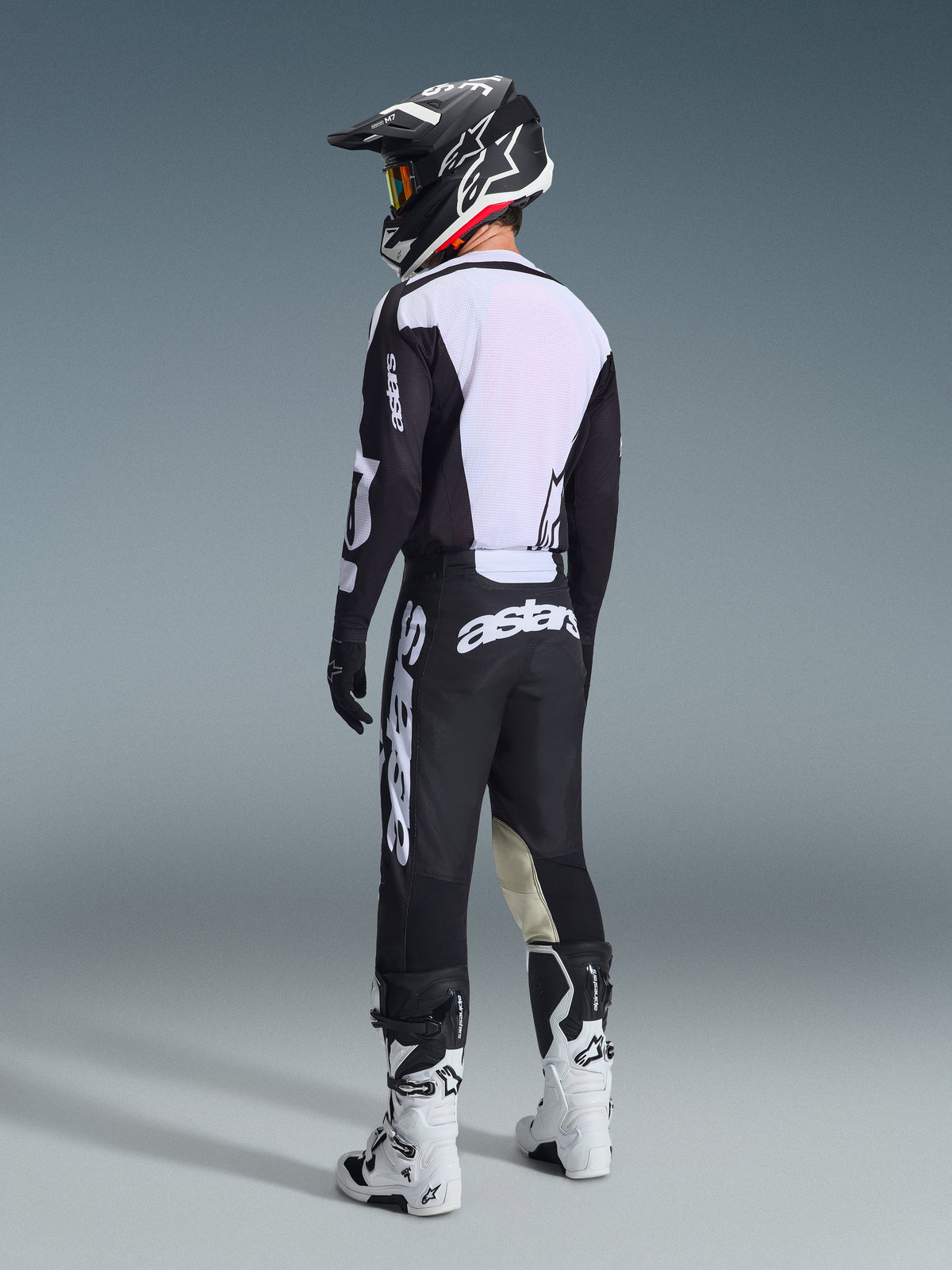 Piloto de motocross visto de trás a usar a Camisola Racer Air Riway 2026 em preto e branco, destacando o grande painel traseiro em malha branca para fluxo de ar, combinada com calças pretas, capacete preto e branco e botas de corrida brancas, equipamento profissional completo para performance todo-o-terreno.