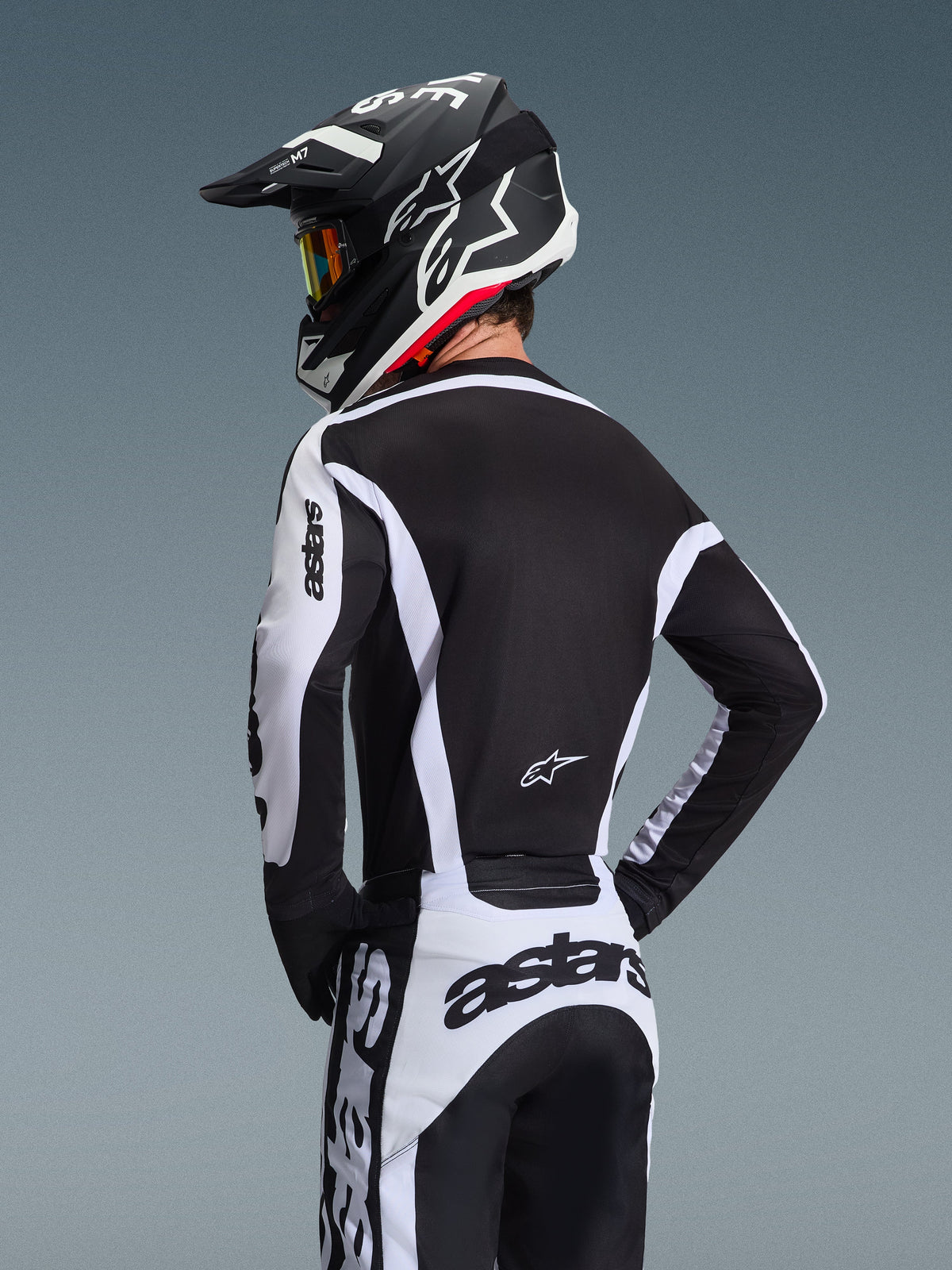 Piloto de motocross visto de trás a usar a Camisola Alpinestars Racer Riway 2026 em branco e preto, com a marca 'astars' nas mangas e na parte inferior das costas, combinada com calças de motocross a condizer e um capacete preto com óculos, equipamento coordenado para corridas todo-o-terreno.