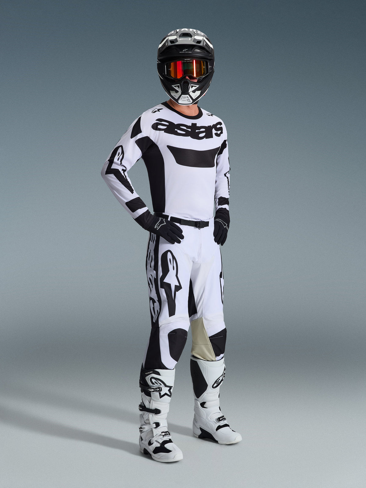 Piloto de motocross com equipamento completo de competição, camisola Alpinestars Racer Riway 2026 em branco e preto, calças de motocross a condizer em branco e preto, capacete preto com óculos espelhados, botas brancas e pretas e luvas pretas, conjunto coordenado para todo-o-terreno