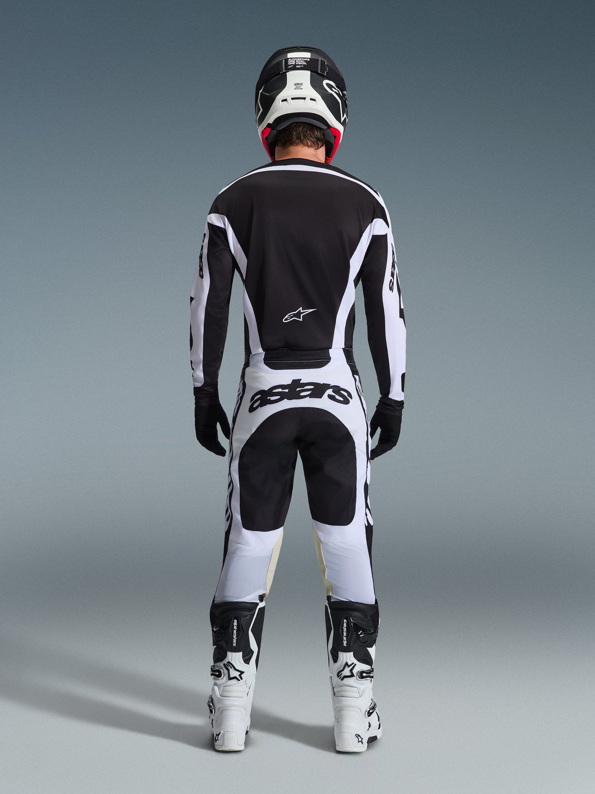 Piloto de motocross de costas com equipamento de corrida completo, Camisola Racer Riway 2026 em branco e preto, calças a condizer com logótipo 'astars' no assento, capacete preto com detalhes brancos, luvas pretas e botas brancas e pretas, equipamento profissional de todo-o-terreno coordenado