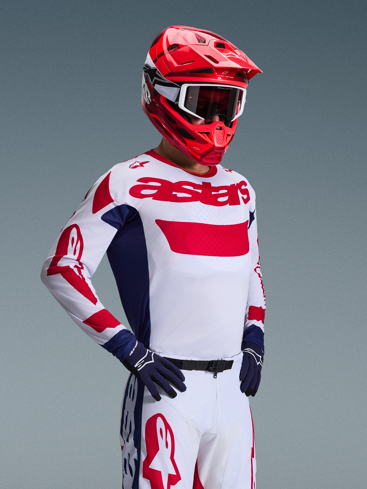 Piloto de motocross a usar a Camisola Alpinestars 2026 Racer Riway em Branco Azul Vermelho, com o logótipo 'astars' a vermelho no peito e painéis laterais azuis, combinada com um capacete vermelho a condizer e calças de corrida brancas para um visual completo de performance off-road.