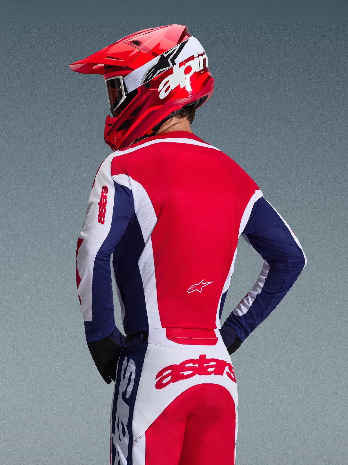 Piloto de motocross visto de costas a usar a Camisola Alpinestars 2026 Racer Riway, em branco, azul e vermelho, calças de off-road a condizer com branding astars marcante, capacete vermelho com logótipo Alpinestars branco, luvas pretas, equipamento de competição de motocross coordenado