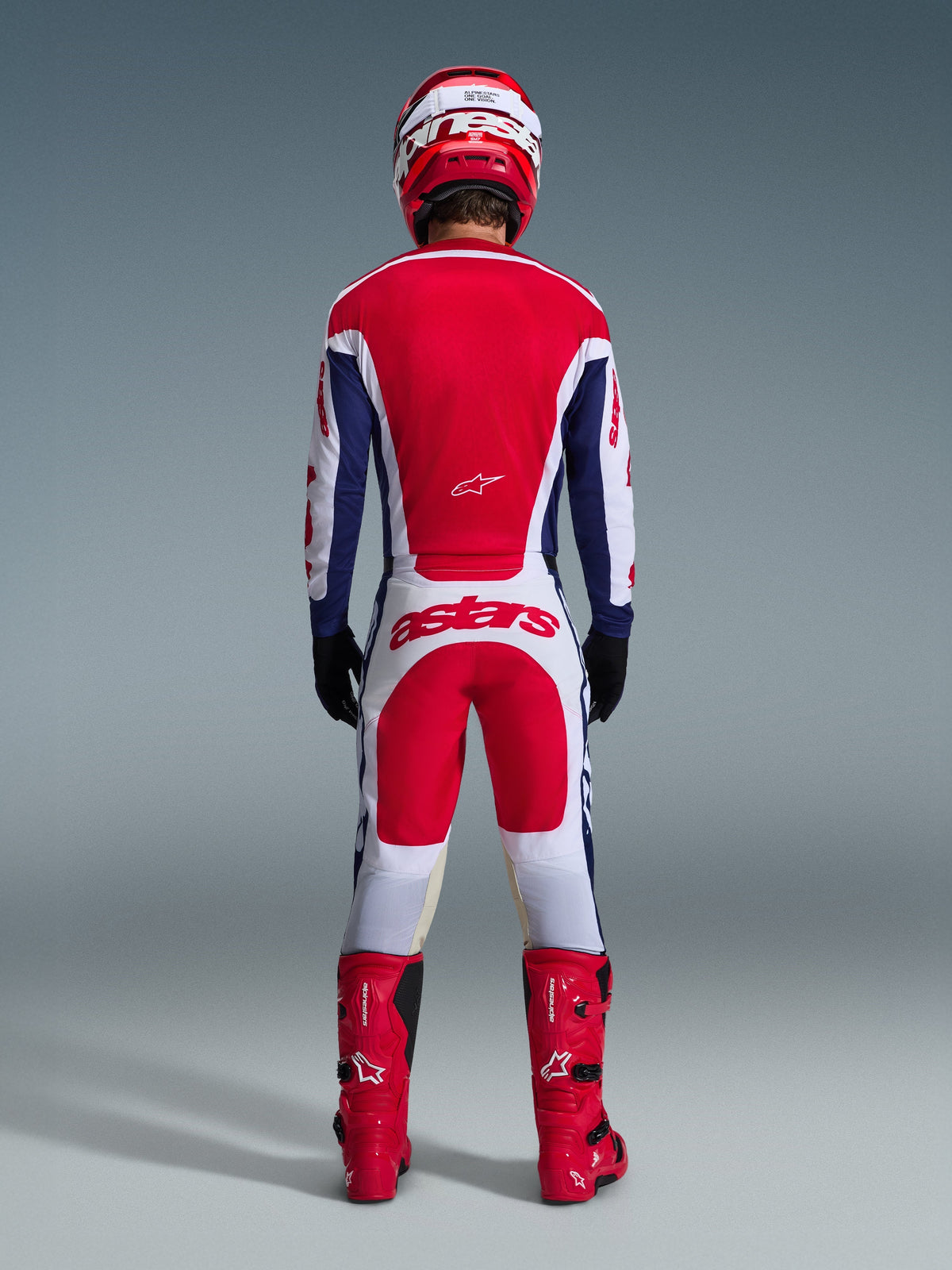 Piloto de motocross com equipamento de corrida completo visto de trás, Camisola Alpinestars 2026 Racer Riway em Branco Azul Vermelho, calças brancas e vermelhas a condizer com a marca 'astars' em destaque, botas vermelhas e capacete vermelho e branco, conjunto de corrida off-road coordenado.