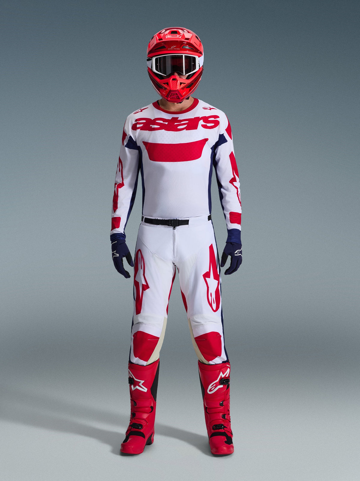 Piloto de motocross com equipamento de corrida completo, Camisola Alpinestars 2026 Racer Riway em branco, azul e vermelho, calças brancas e vermelhas a condizer, capacete vermelho, botas vermelhas e luvas azul-marinho, conjunto coordenado para todo-o-terreno