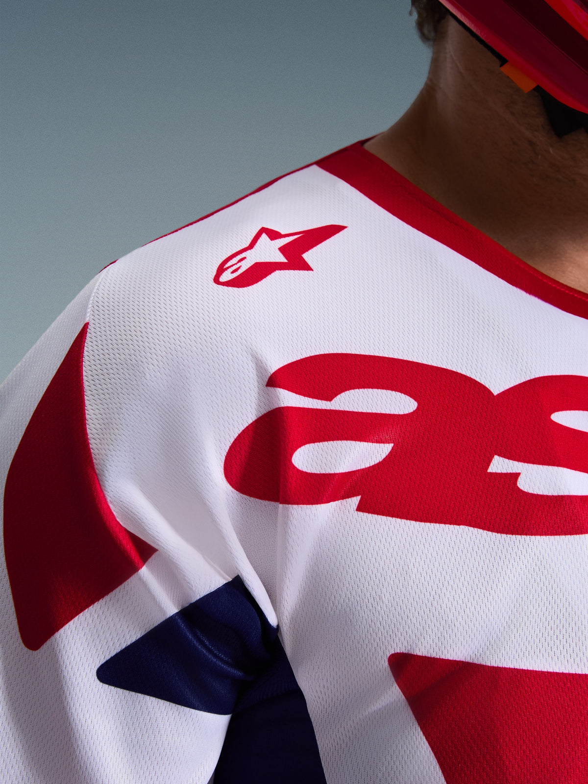 Pormenor da Camisola Alpinestars 2026 Racer Riway, camisola de motocross em Branco, Azul e Vermelho, com textura de malha respirável que absorve a humidade, logótipo de estrela vermelha no ombro e gráficos vermelhos arrojados no peito