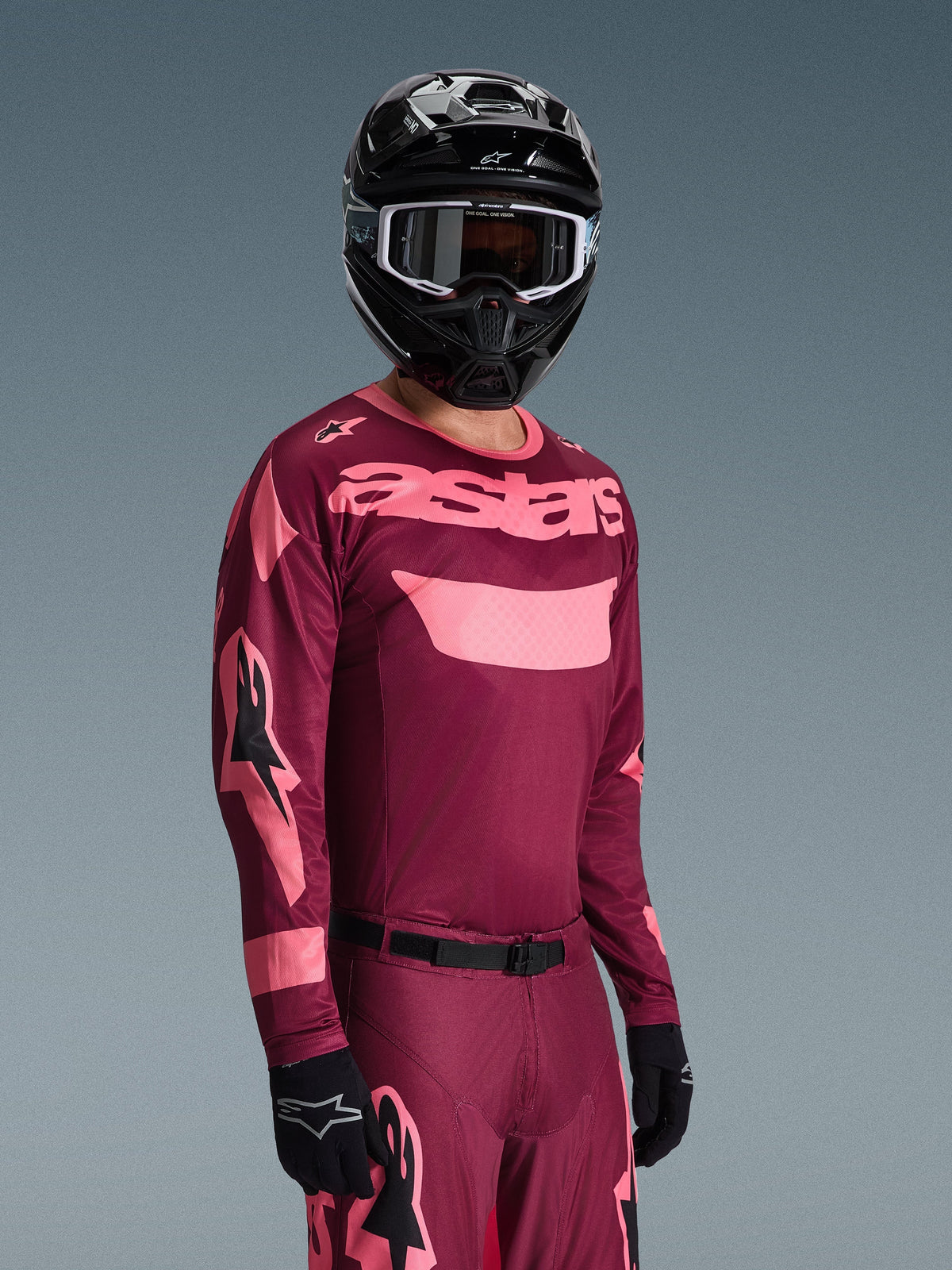 Piloto de motocross a usar a camisola Alpinestars 2026 Racer Riway em Dark Red Coral com calças a condizer, com a marca 'astars' em cor-de-rosa coral no peito, acompanhado de capacete e luvas pretas, conjunto de equipamento de corrida todo-o-terreno.