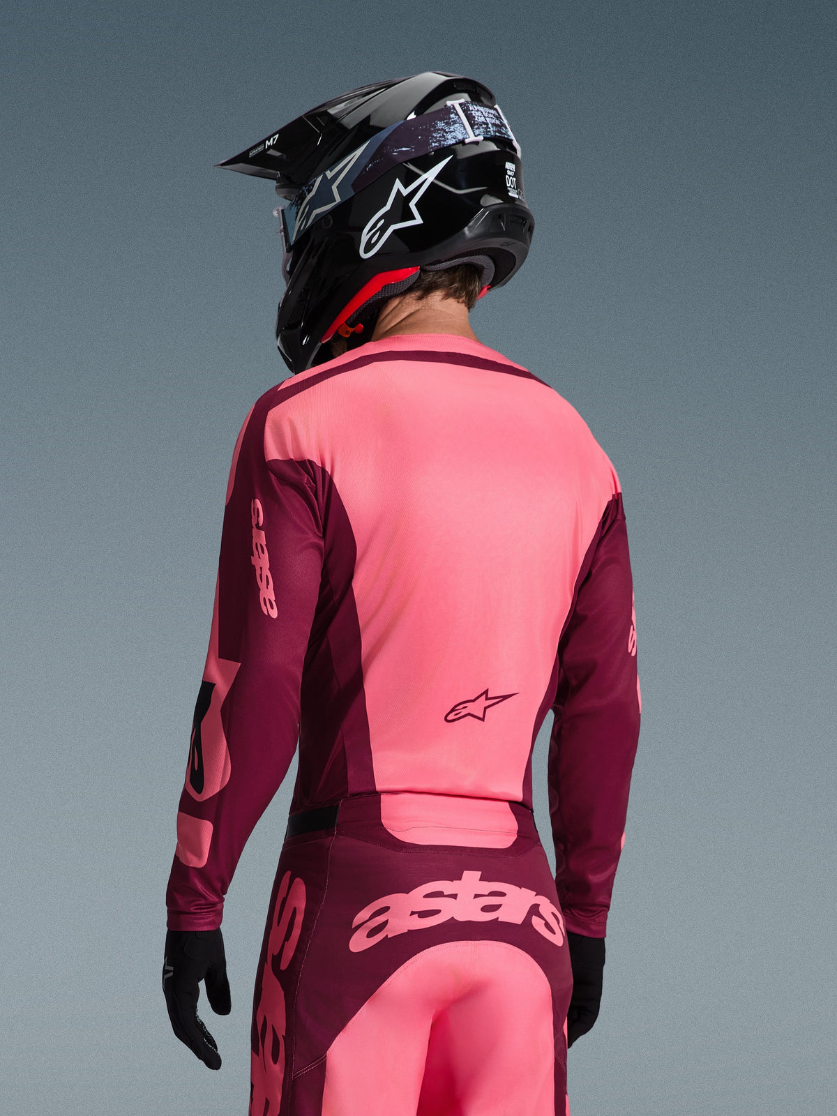Piloto de motocross visto de costas a usar a camisola Alpinestars Racer Riway 2026 e calças a condizer, combinação de cores vermelho escuro e coral, capacete preto com logótipo Alpinestars branco, luvas pretas, equipamento profissional coordenado para todo-o-terreno