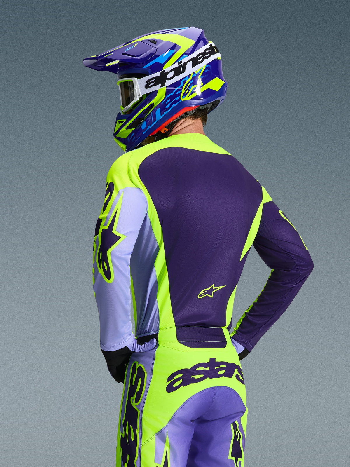 Piloto de motocross visto de trás a usar a camisola Alpinestars 2026 Racer Portl e as calças a condizer, esquema de cores roxo e amarelo fluo com detalhes em lavanda, capacete coordenado azul e amarelo, equipamento com ajuste de corrida concebido para desempenho todo-o-terreno