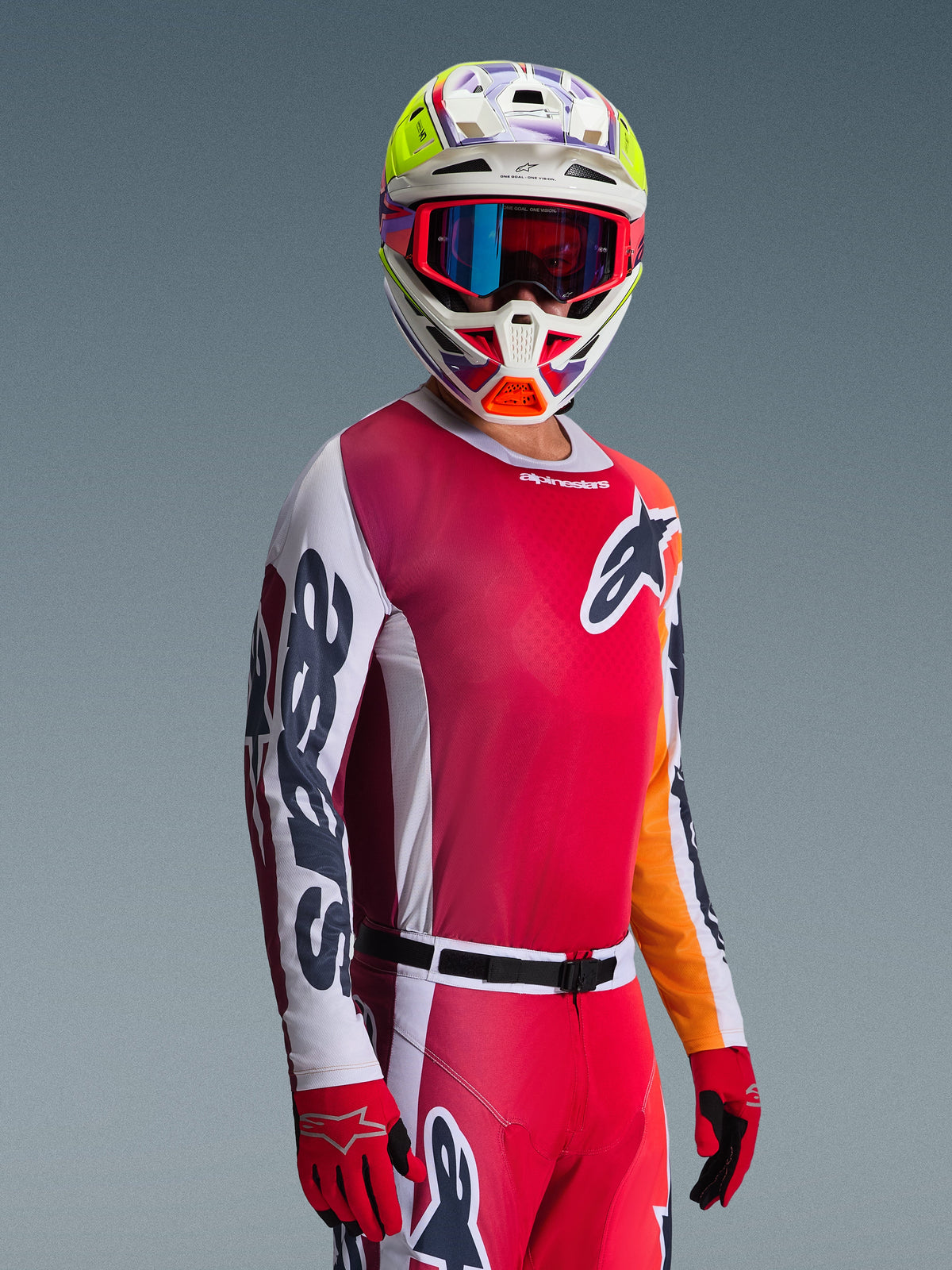 Piloto de motocross a usar o conjunto de equipamento Alpinestars 2026 Racer Portl, com camisola vermelha de tons rosa e mangas brancas, calças vermelhas a condizer, capacete branco com detalhes vibrantes em amarelo e roxo, óculos vermelhos com lentes azuis iridescentes e luvas vermelhas, equipamento de corrida profissional.