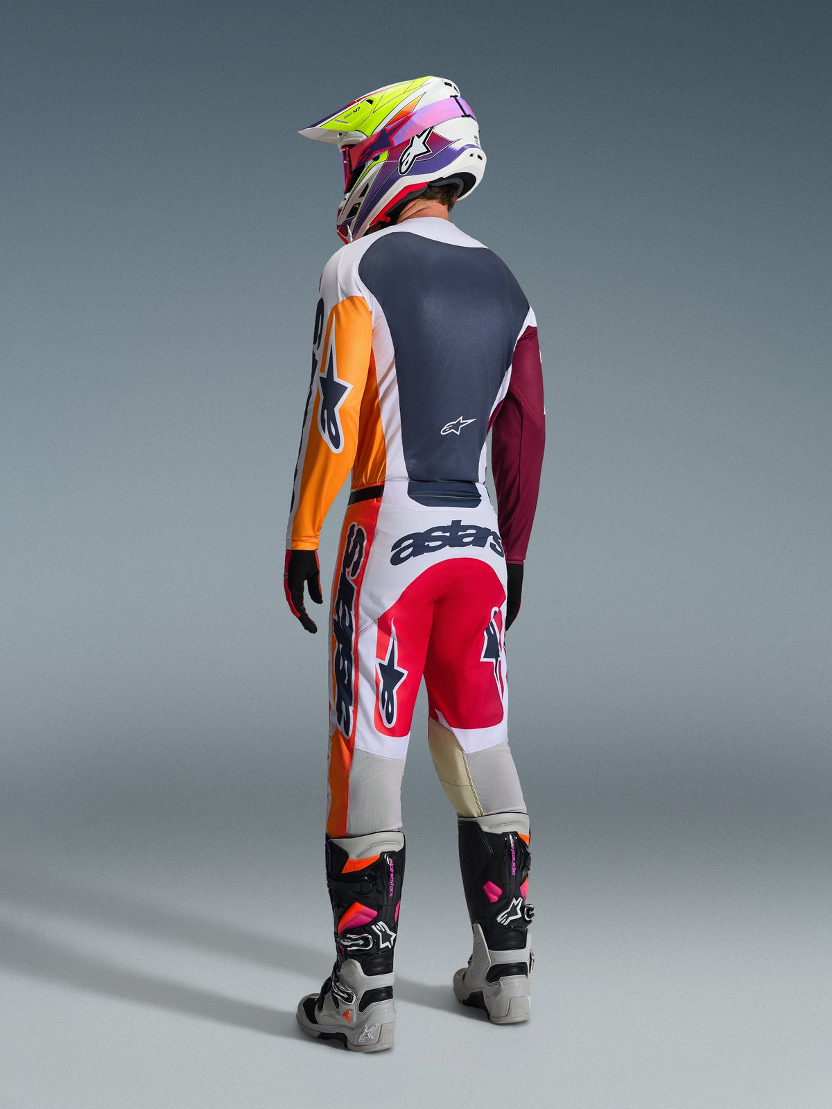 Piloto de motocross visto de trás a usar o Conjunto de Equipamento Alpinestars Racer Portl 2026, com camisola em blocos de cor com mangas cor de laranja e bordeaux, calças brancas com painéis vermelhos e marca 'astars' em destaque, capacete multicolorido e botas cinzentas, fato de competição off-road profissional.