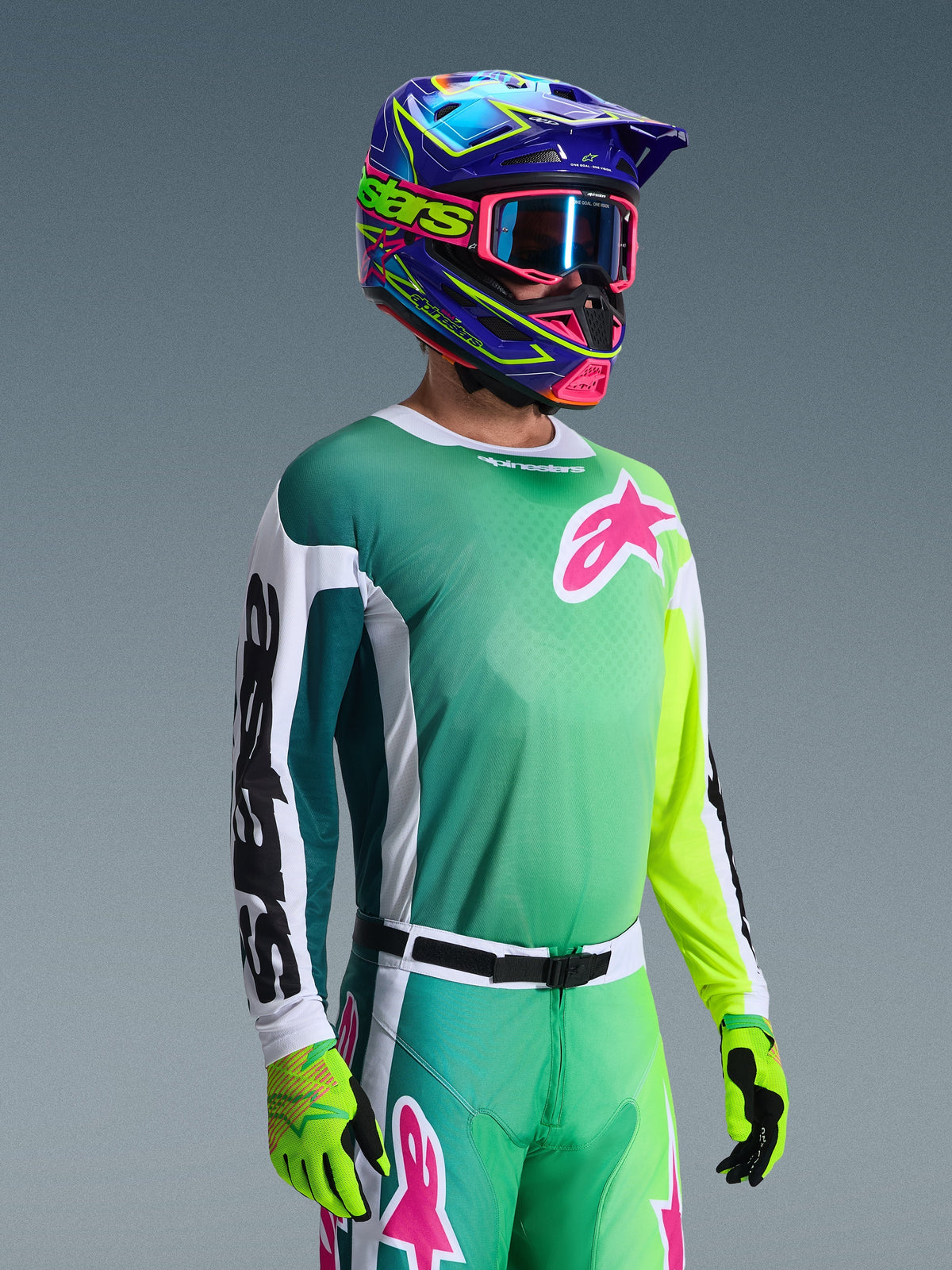 Alpinestars 2026 Racer Portl Jersey, camisola de motocross, verde, branca e preta, usada por um piloto com calças verdes a condizer, luvas fluorescentes e um capacete azul e roxo vibrante com óculos cor-de-rosa, apresentando um logótipo Astars cor-de-rosa no peito