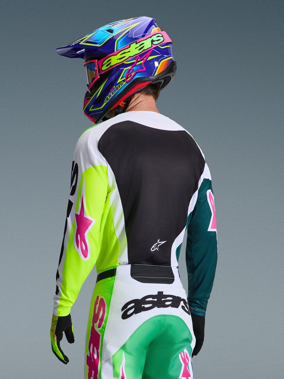 Piloto de motocross visto de costas com a camisola Alpinestars 2026 Racer Portl em Green White Black, com logótipos de estrela cor-de-rosa, combinada com calças a condizer e um capacete colorido azul e roxo, equipamento de corrida coordenado para performance off-road.