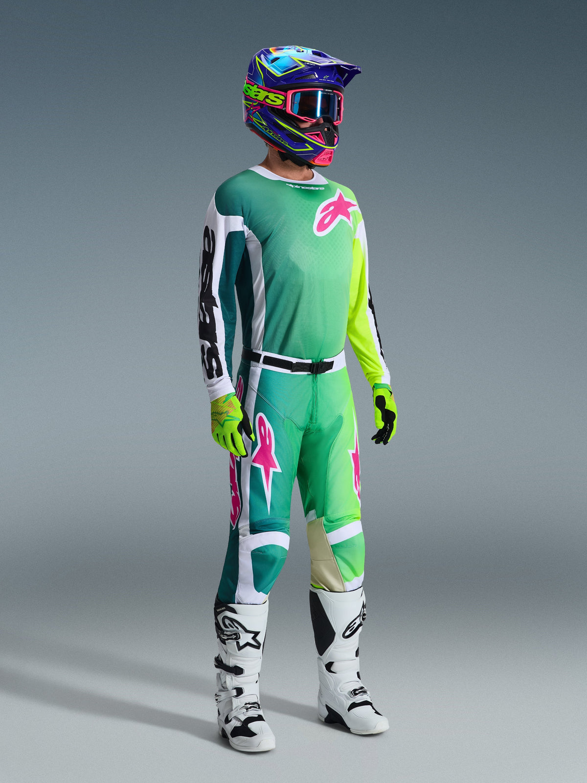 Piloto de motocross com equipamento de competição completo, Camisola Racer Portl 2026 em verde com mangas brancas e pretas e logótipo cor-de-rosa, calças verdes a condizer com detalhes cor-de-rosa, capacete multicolorido com óculos, botas brancas e pretas e luvas verdes, conjunto coordenado para corridas off-road