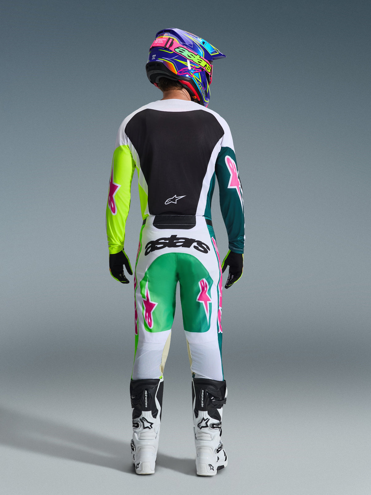 Vista traseira de um piloto de motocross a usar o conjunto de equipamento Alpinestars 2026 Racer Portl, camisola branca com painel traseiro preto e mangas em amarelo néon e verde escuro com estrelas cor-de-rosa, calças brancas com detalhes em verde e verde-azulado e logótipos de estrelas cor-de-rosa, acompanhado por capacete colorido, luvas pretas e botas brancas e pretas.