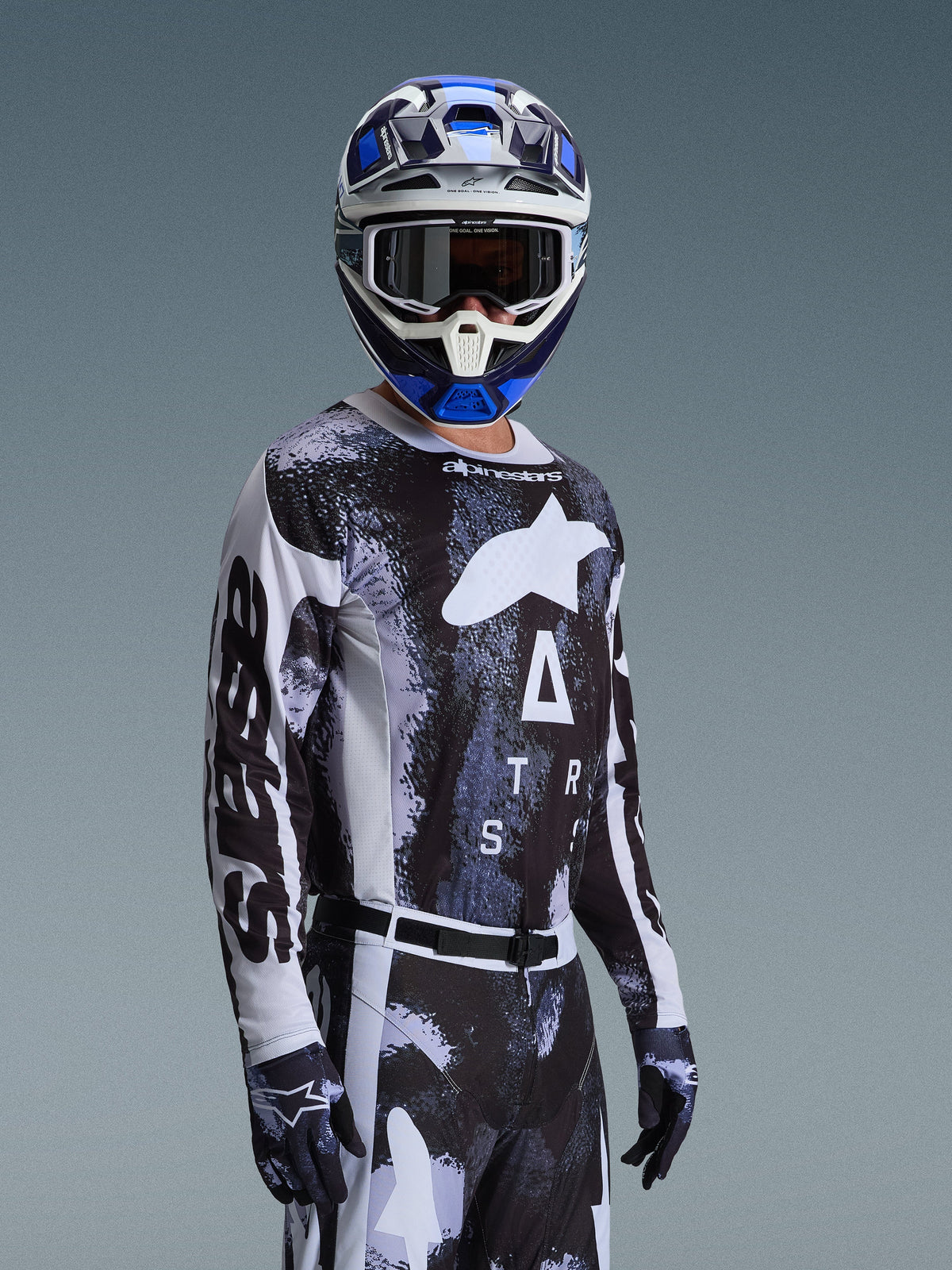 Piloto de motocross a usar a Camisola Alpinestars 2026 Racer Lahnd em Iron Camo, com um padrão de camuflagem em preto e cinzento e um grande logótipo Alpinestars branco no peito, combinada com um capacete e luvas a condizer para corridas fora de estrada.