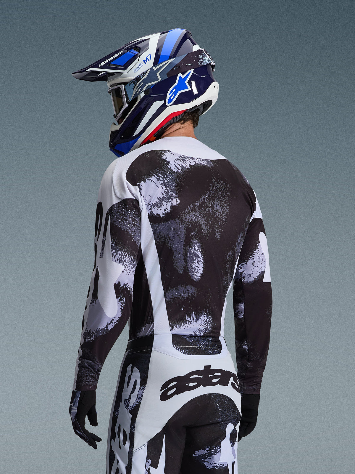 Camisola Alpinestars Racer Lahnd 2026 em Iron Camo, vista traseira de um piloto de motocross com conjunto de equipamento de padrão abstrato preto, branco e cinzento, incluindo calças a condizer, luvas pretas e capacete de corrida azul com óculos de proteção.