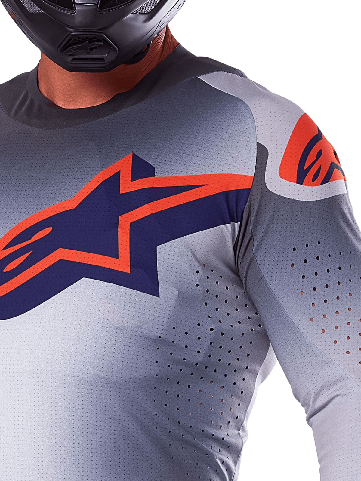 Pormenor de um piloto com a Camisola Alpinestars 2025 Supertech Lipan, camisola de motocross, cinza e laranja fluo, com um grande logótipo Alpinestars laranja no peito, orifícios de ventilação cortados a laser e um capacete preto, equipamento de competição profissional.