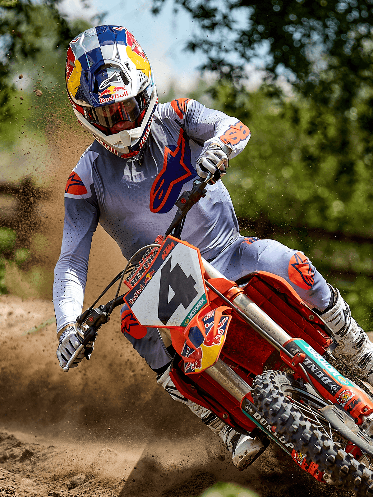 Piloto de motocross em ação numa pista de terra, a usar a Camisola Alpinestars 2025 Supertech Lipan em Cinza Laranja Fluo, calças cinzentas com detalhes laranja, botas brancas e capacete com a marca Red Bull, a curvar numa mota de motocross laranja com projeção de terra, equipamento profissional de corrida off-road