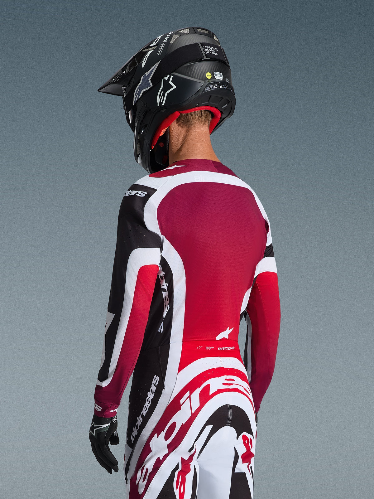 Vista traseira de um piloto de motocross com a camisola Supertech AFD Vista 2026 em preto, vermelho e branco, combinada com calças a condizer e um capacete preto, apresentando logótipos Alpinestars em destaque, painéis de malha vermelhos e um corte atlético de competição.