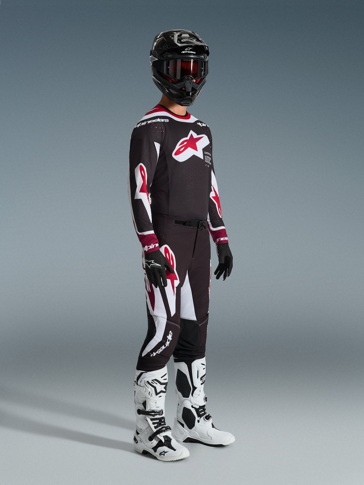 Piloto de motocross com equipamento de corrida completo, camisola Alpinestars 2026 Supertech AFD Vista em preto, vermelho e branco, calças de motocross a condizer, capacete preto com óculos vermelhos, botas brancas e pretas e luvas pretas e brancas, conjunto coordenado para competição off-road profissional